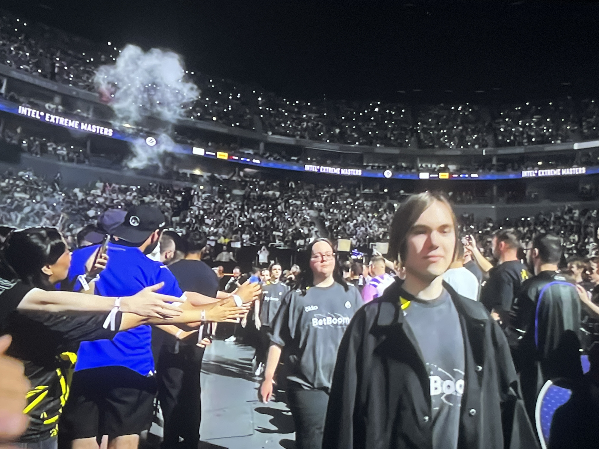 The image depicts a nighttime scene at a large indoor event, possibly an esports tournament, with a roaring crowd filling stadium seating in the background. The word "INTEL" is clearly visible on banners hanging in the stadium, along with "EXTREME MASTERS" in multiple locations. Small lights are being held up by spectators, creating a starry effect in the stands.
In the foreground, a group of people are walking on a dark surface, perhaps a stage or walkway. A woman in a black shirt with yellow accents and a man in a bright blue shirt and black shorts are visible on the left, interacting with someone off-camera. In the center, a woman wearing glasses and a grey t-shirt with "BetBoom" written on it, walks forward. To her right, a young person with long hair, wearing a grey t-shirt with "BetBoom" visible and a black jacket, is also walking forward and looking towards the viewer. Other individuals, some in black shirts, are interspersed in the crowd behind them.
A large plume of white smoke or fog is seen in the upper left quadrant of the image, adding a dramatic element to the atmosphere. The lighting suggests an indoor stadium setting, with spotlights and the ambient light from the crowd and stage creating a dynamic visual. The presence of the "BetBoom" logo on multiple shirts indicates that this event is sponsored by or features this organization. The overall mood appears to be exciting and energetic, typical of a live competitive event.