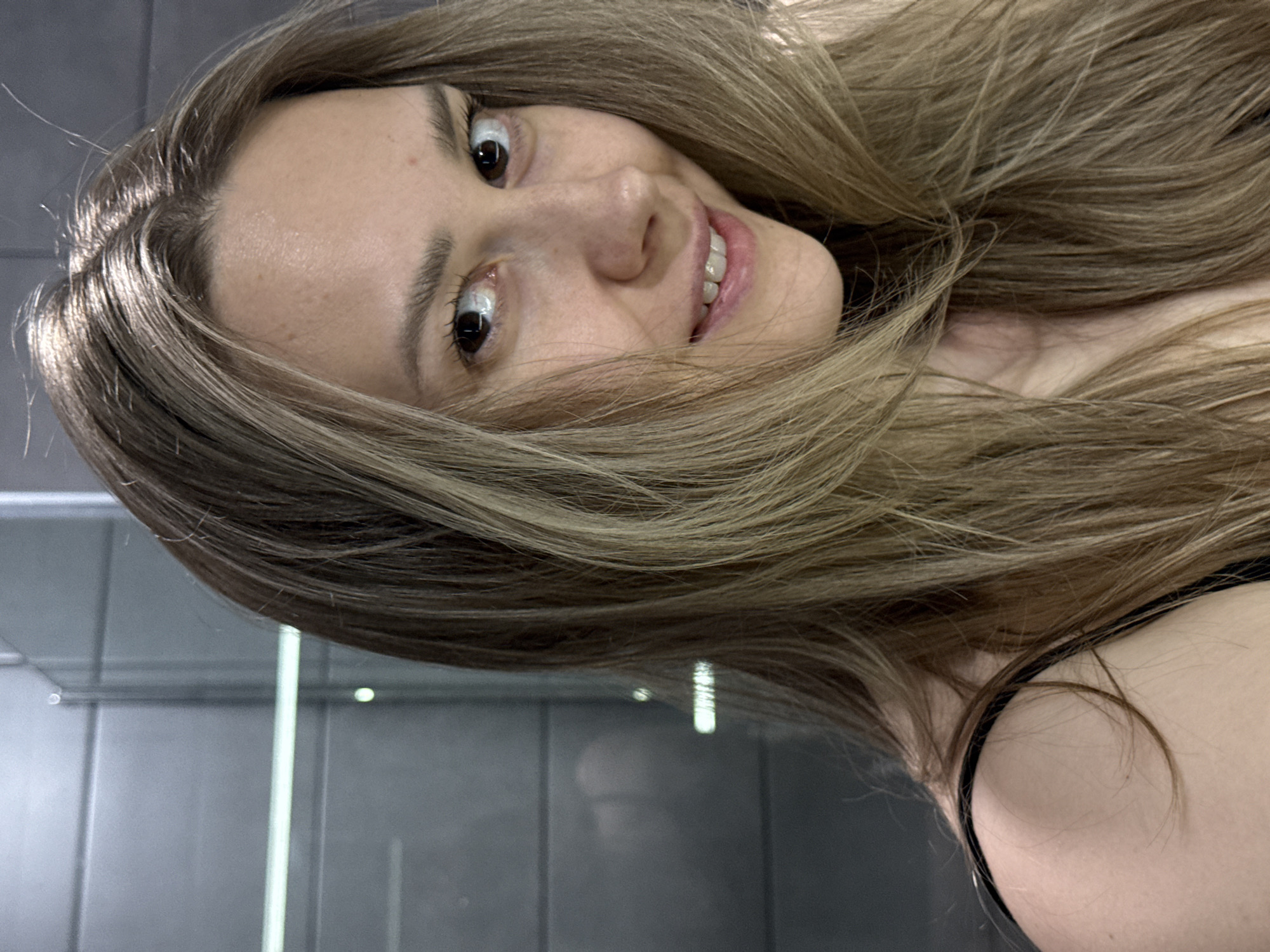 A young woman with long, light brown hair is smiling at the camera. Her hair is partially obscuring her face. She is wearing a black top. The background appears to be a bathroom with grey tiled walls and a shower. The scene is possibly located in an indoor setting. There is no information available to ascertain the location to be in Chaikovsky, Russia.