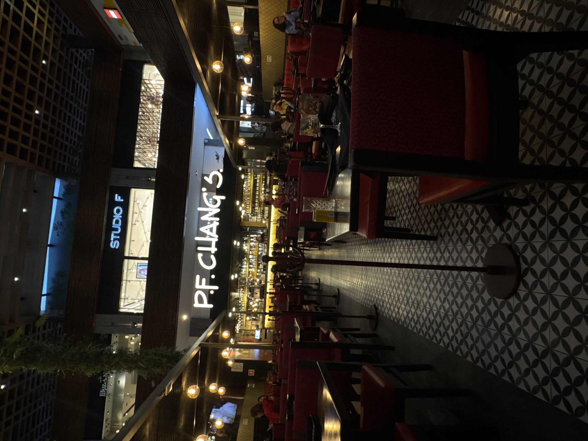 This image depicts the interior of a P.F. Chang's restaurant, situated within the Angelópolis Lifestyle Center shopping mall in Puebla, Mexico. The perspective is from the dining area, looking towards the restaurant's bar and beyond into the mall's multi-level common space.
The restaurant's dining area features numerous dark rectangular tables paired with red upholstered chairs and banquettes. The floor is covered in black and white patterned tiles displaying a repeating circular or quatrefoil motif. Spherical pendant lights with a warm glow hang from an overhead dark, slatted wooden ceiling structure. A tall, dark metal coat rack stands prominently in the central aisle.
Approximately 10-15 individuals are present within the restaurant. Several patrons are seated at tables on both sides of the central aisle. For instance, a person in a red top is visible seated on the left, and another individual in a blue top is visible on the far right. Indistinct figures are also visible near the illuminated bar at the rear of the restaurant.
Beyond the restaurant's immediate confines, elements of the shopping mall are visible. Prominent illuminated signs for "STUDIO F" and "BIRAN" are visible on an upper level. A large vertical column wrapped in green foliage descends from an upper level. The mall's architecture includes dark wooden paneling and an open, grid-like ceiling structure. A "LEGO" logo is partially visible on the upper right. The scene is illuminated by a combination of the restaurant's warm, dim lighting and the brighter, cooler light emanating from the mall's storefronts and general ambient lighting.