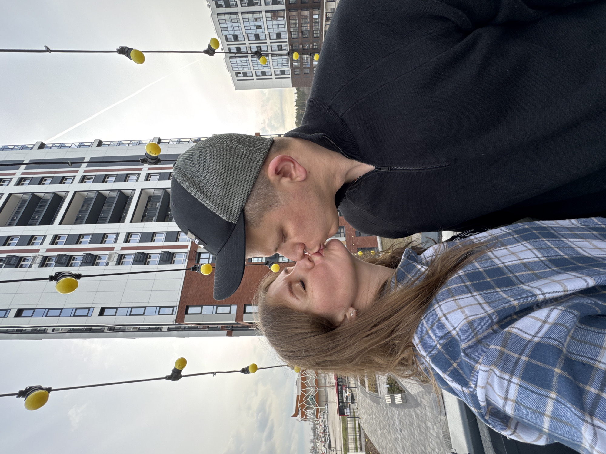 A couple is kissing on an elevated walkway in Perm, Russia. The woman wears a blue and white plaid shirt. The man wears a black jacket and a dark baseball cap. In the background are buildings and string lights with yellow bulbs. A parking lot with cars is visible below. The sky is overcast.
