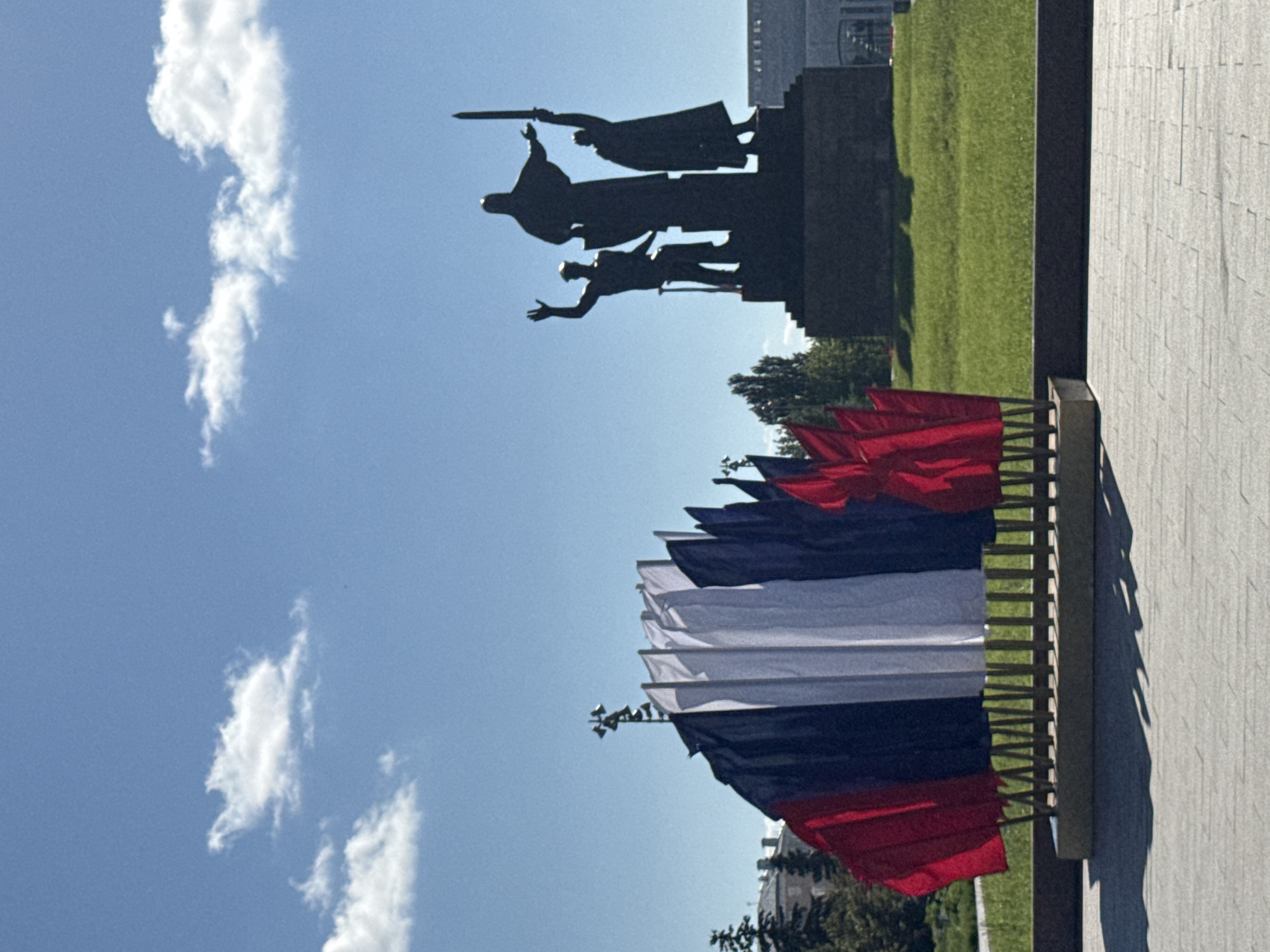 A cluster of small flags, arranged in a fan shape with vertical stripes of red, white, and blue, is displayed in the foreground. Behind the flags, a silhouetted monument features three figures standing on a pedestal; the central figure is taller than the other two, and the figure on the right holds a sword aloft. The monument is situated on a grassy area. The sky above is blue with scattered white clouds. The scene appears to be outdoors, likely in a public space such as a park or plaza. This is a photograph of the Monument of Glory on the esplanade of the Kama River embankment in the city of Perm, Russia.