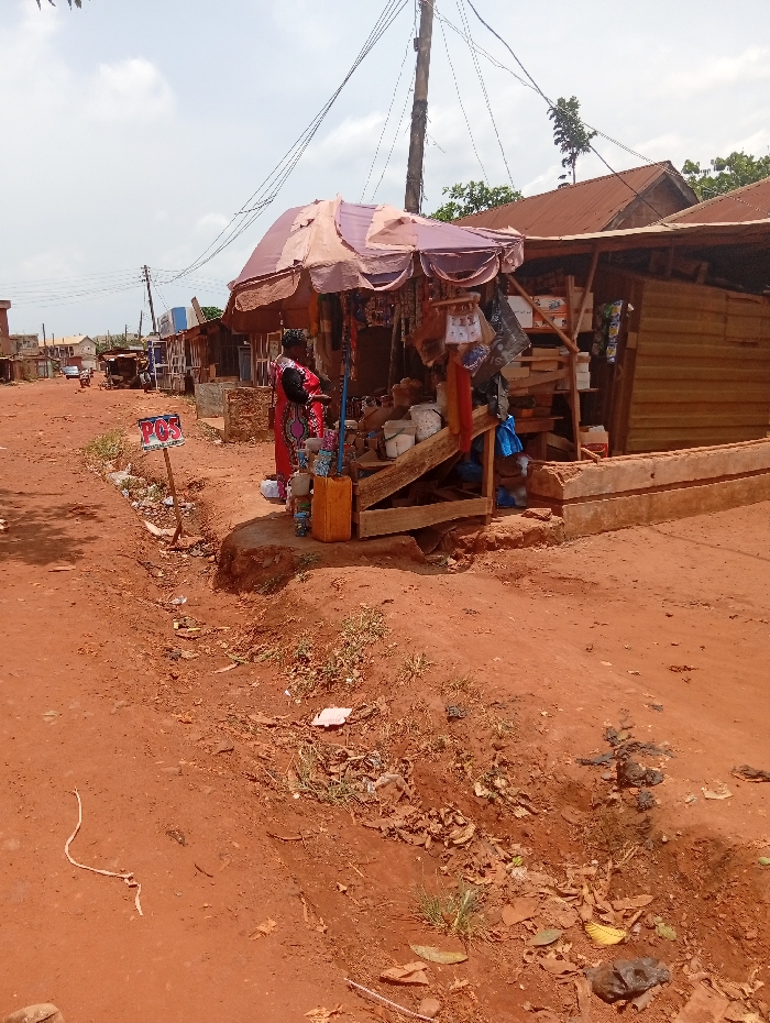The scene depicts a roadside commercial area in Nsukka, Nigeria, under a bright, overcast sky. A wide, red-brown dirt road, indicative of laterite soil, dominates the foreground and extends into the distance. The road's edge features a shallow, eroded ditch containing sparse green vegetation, dry leaves, and scattered refuse including plastic packaging and paper.
In the mid-ground, on the right, a rudimentary retail stall stands elevated on an earthen platform. The stall is constructed from a wooden frame, partially enclosed with corrugated metal or wooden planks, and shaded by a faded pink-brown umbrella. Various consumer goods, including bags, plastic items, and packaged products, are displayed hanging from the umbrella structure and arranged on wooden shelves within the stall. A yellow plastic jerrycan rests on the ground beside the stall.
A female individual, dressed in a patterned red and black gown, stands facing the goods at the stall, appearing to observe or interact with the merchandise. She carries a dark shoulder bag. Behind the stall, a tall utility pole with numerous overhead power lines rises against the sky. Further down the road, a series of similar, simple wooden structures with reddish corrugated metal roofs line the roadside, indicating additional shops or dwellings.
To the left of the main stall, near the roadside, a white sign with "POS" printed in red text is attached to a wooden stake. In the far distance along the dirt road, a light-colored vehicle and a small, dark animal (possibly a chicken or dog) are visible.