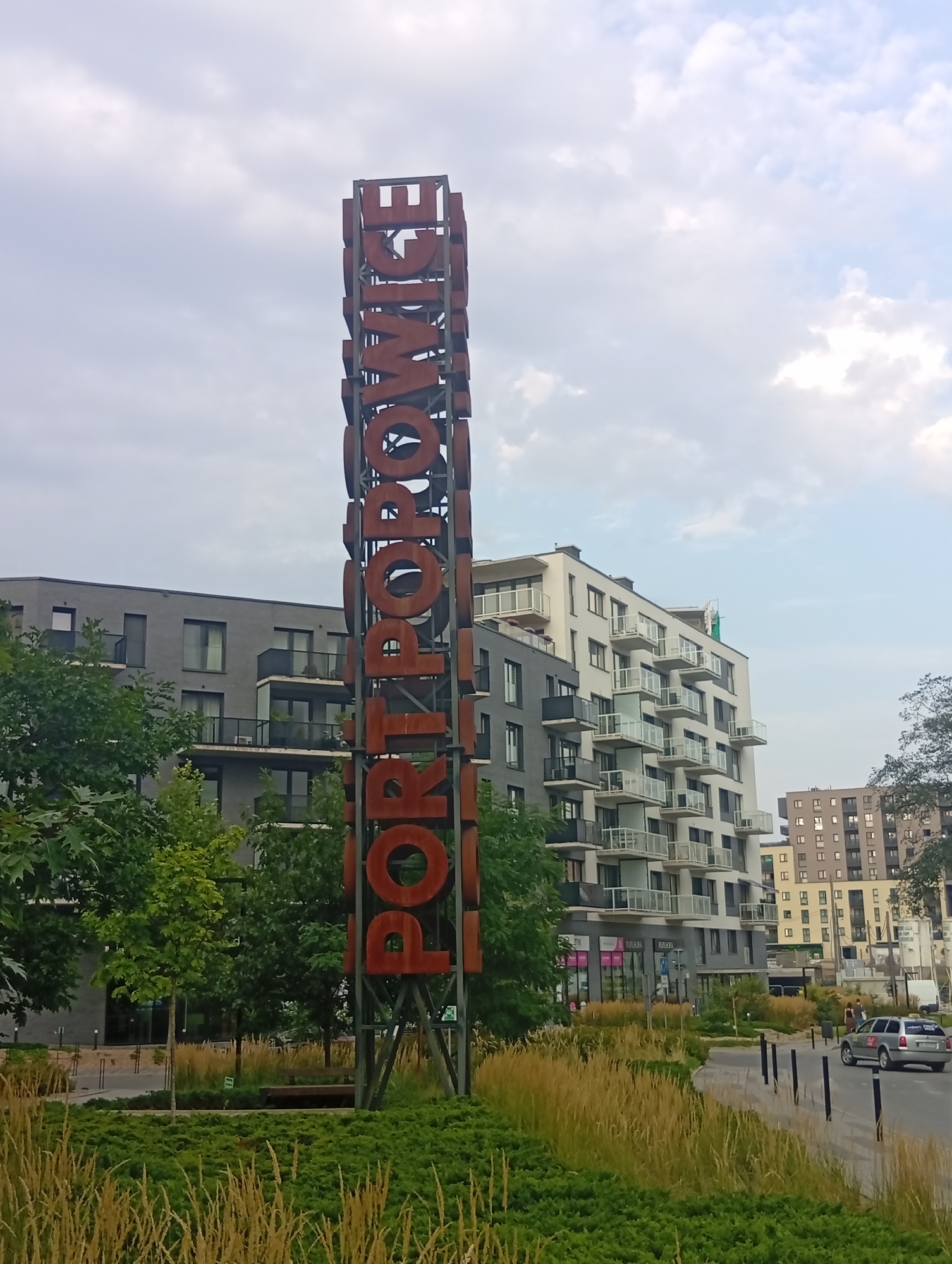 An outdoor, eye-level, slightly tilted full shot captures a tall, rust-colored metal sculpture with large, cutout letters spelling "PORTOWICE" against a cloudy sky. The sculpture is positioned in the foreground, to the right of center. Behind the sculpture, on the left, is a modern, grey apartment building with dark-framed windows and balconies. To the right of that, a white apartment building with numerous balconies extends upward. Further in the background, another residential building with lighter-colored facade and many windows is visible. A paved pathway curves through a landscape of green bushes and tall, dry grasses in front of the buildings. A silver car is seen driving away on the right side of the road, with two pedestrians walking on the sidewalk in the distance. The scene appears to be a contemporary urban development in Wrocław, Poland, likely a residential area.