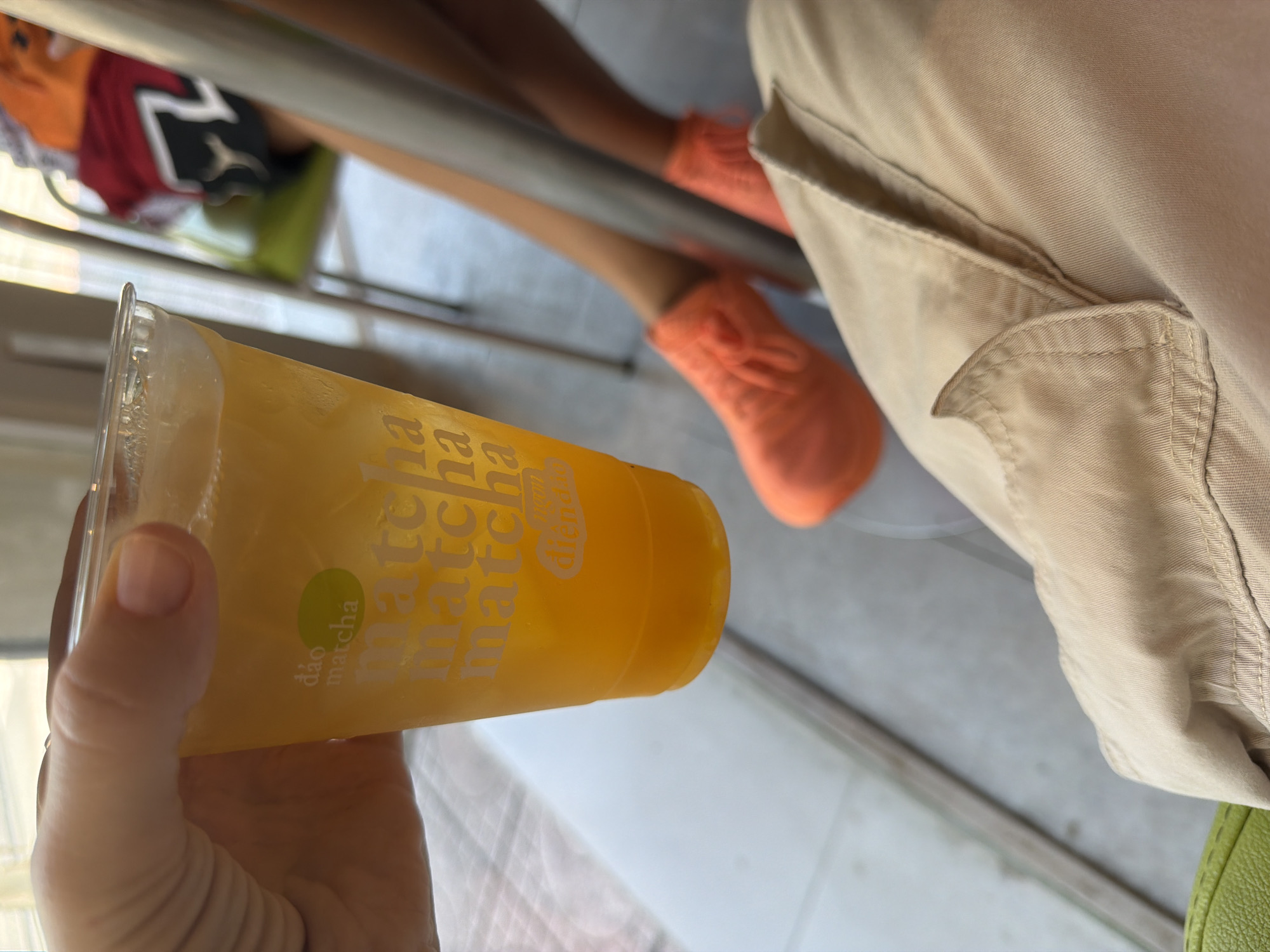 A first-person perspective shot shows a hand holding a clear plastic cup filled with an orange beverage and ice. The cup has white text on it, including "dao matcha" and multiple instances of "matcha." The background is blurred, showing parts of a seated person wearing orange shoes and a chair. The scene appears to be an indoor establishment, possibly a cafe or restaurant, with natural light filtering in. The beverage is being held up and tilted slightly forward, indicating it might be offered or presented. The setting is likely in Nha Trang Ward, Vietnam, given the context provided.