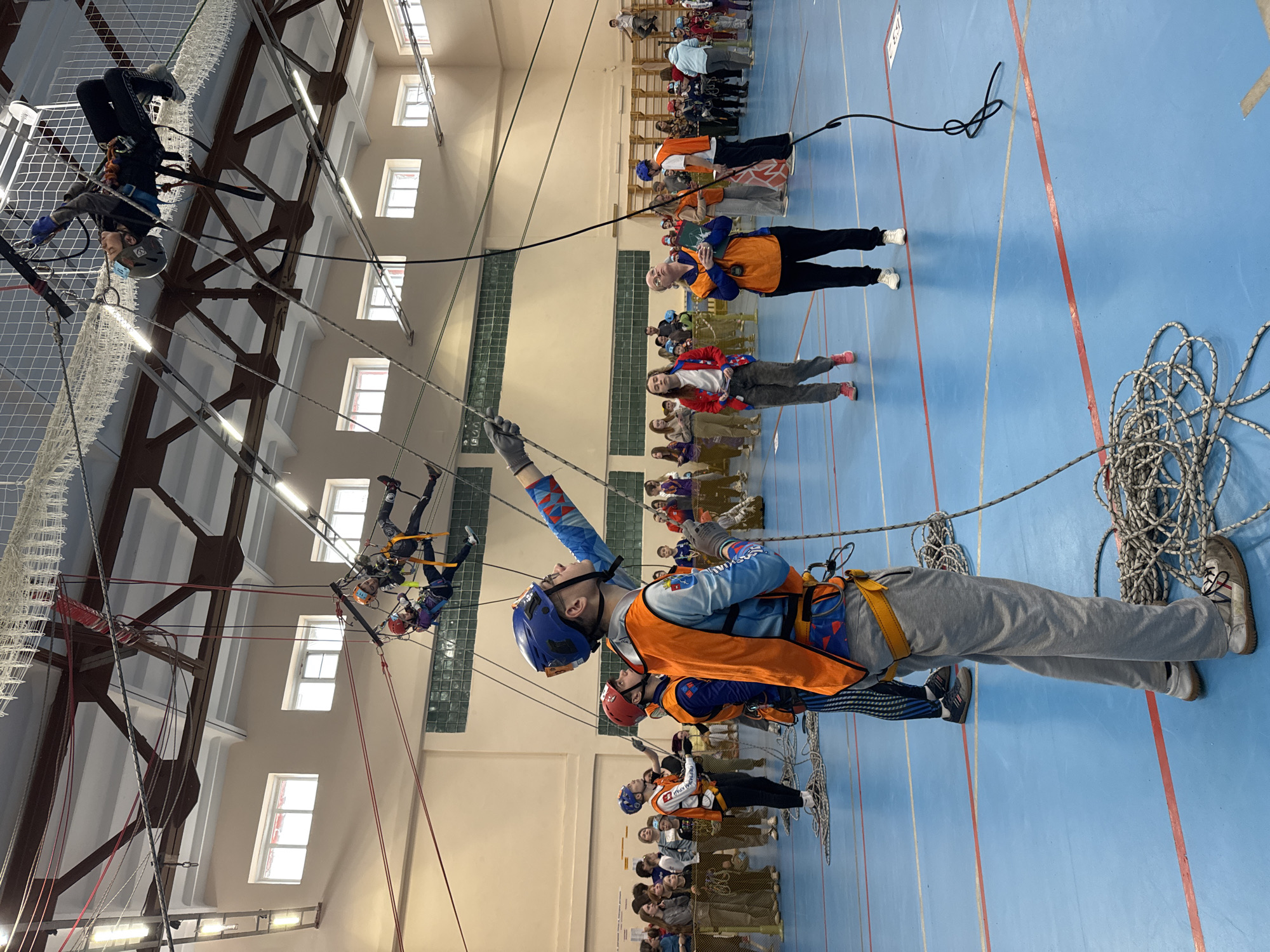 The image shows an indoor climbing activity in a large hall, possibly in Orel, Russia. Several individuals are secured with ropes and harnesses, suspended from the ceiling's metal framework. One person is upside down, while others are at various heights. On the floor, a group of people stand and watch, some wearing vests and helmets, indicating they might be instructors or participants. A coiled rope lies on the blue floor. The hall has high ceilings with multiple windows and rows of fluorescent lights.