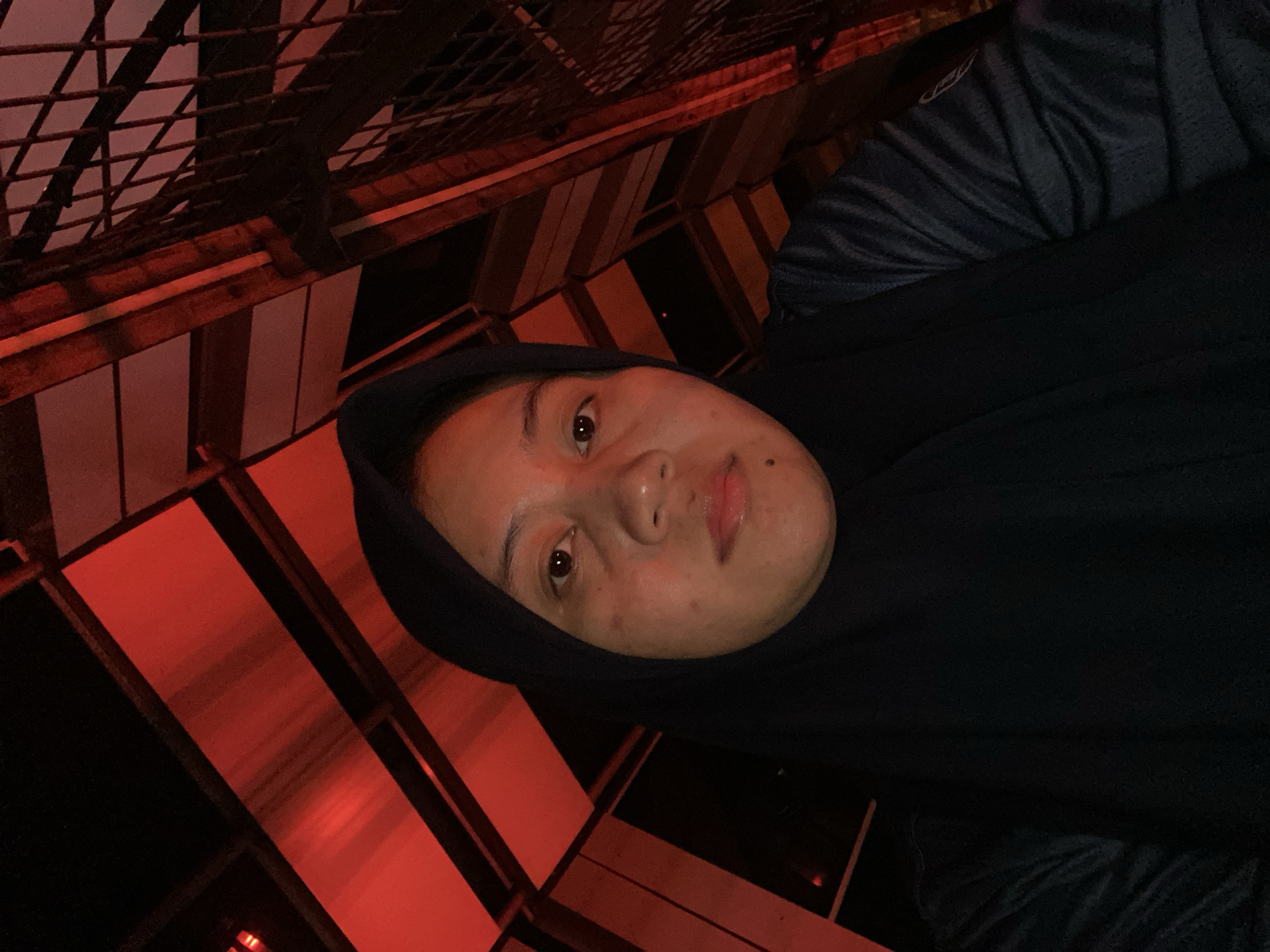 A selfie is taken from a low angle of a young woman wearing a black hijab and a dark blue shirt. She is looking directly at the camera, and her face is partially illuminated by red lighting. The background is a complex structure of intersecting beams and panels, bathed in red light, suggesting an indoor or covered outdoor space with architectural features. The lighting is dramatic, with strong red hues casting shadows and highlighting textures. The woman's skin appears to have some blemishes. The environment has a modern, possibly industrial or artistic aesthetic, with geometric patterns formed by the structure. The location is identified as Banjarbaru, Indonesia.