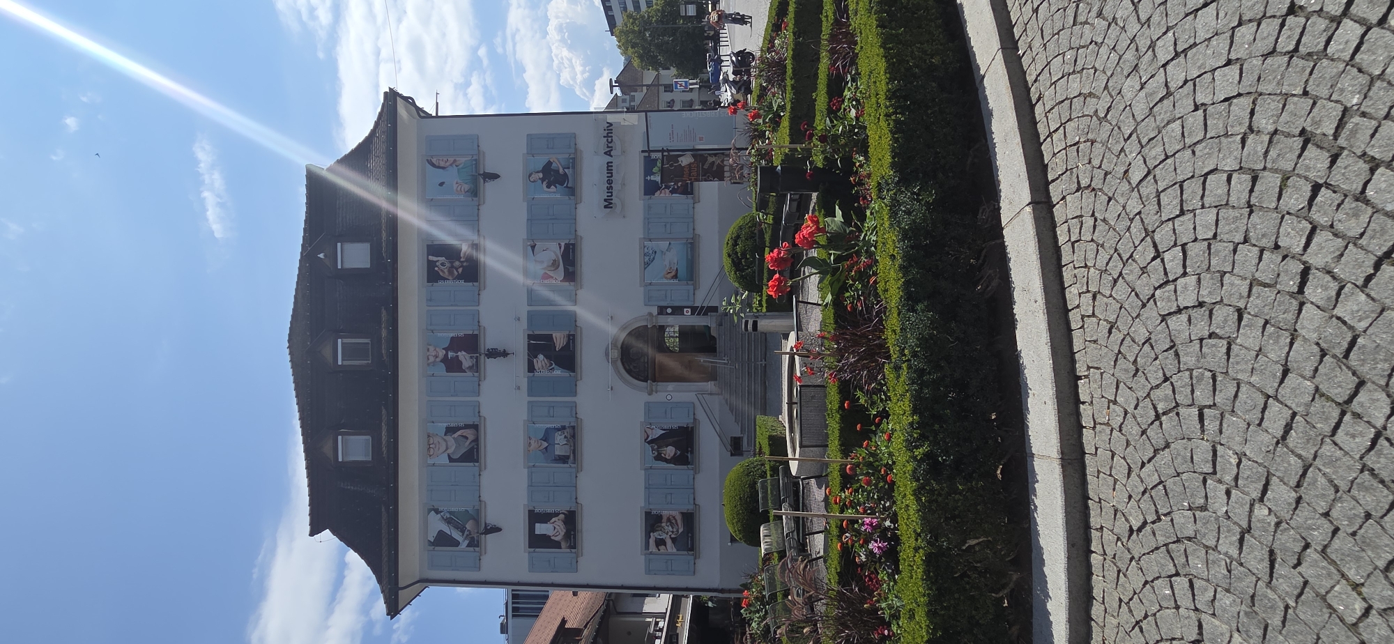 The image shows the exterior of a building, identified as the Museum Archiv in Dornbirn, Austria. The building has a light-colored facade, a dark mansard roof with dormer windows, and blue shutters on its windows. Each window displays a printed image of a person holding an object or engaging in an activity. In front of the museum is a garden area with manicured hedges, bushes, red flowers, and dark ornamental grasses. A circular stone fountain is visible in the garden. The foreground is a cobblestone street. The sky is blue with scattered clouds and a strong sun glare is present in the upper right portion of the image. In the background, other buildings and trees are visible. A person on a bicycle is seen on the right side of the image.
