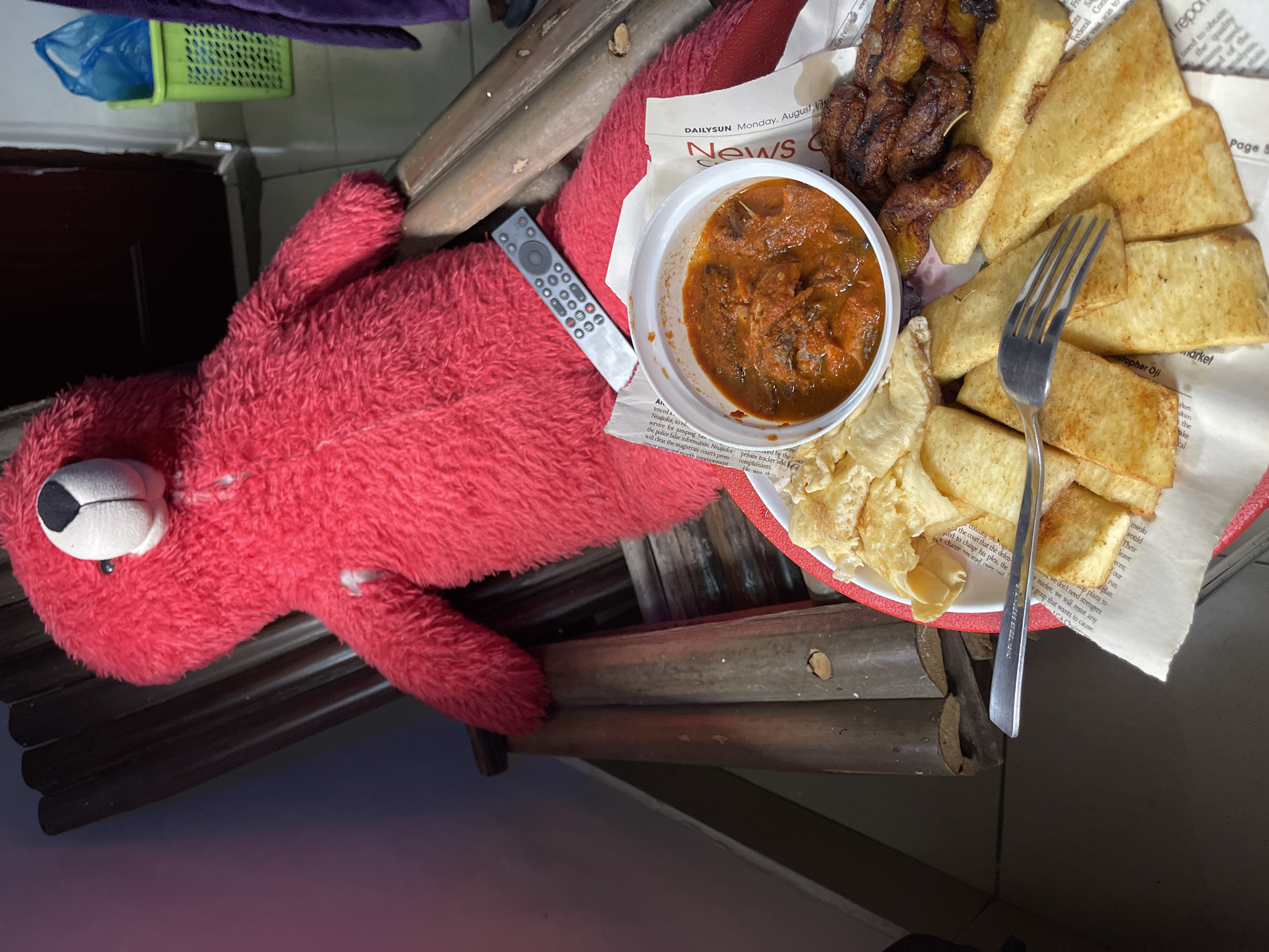 A photograph shows a red, plush Elmo toy lying horizontally on a wooden surface. A remote control is placed alongside the Elmo toy. Next to Elmo is a white plate lined with newspaper and containing fried yams, fried plantains, and a white bowl with a stew-like substance. A fork is on the plate. The newspaper is identifiable as "DAILY SUN Monday, August", placing the scene on or after that date. The partial text visible, along with the mention of Nnajiofor, suggests a location in Nigeria. Gwarinpa is specified as a possible location within Nigeria. The surrounding environment is partially visible, including a green basket, and dark furniture. The scene appears to be indoors.