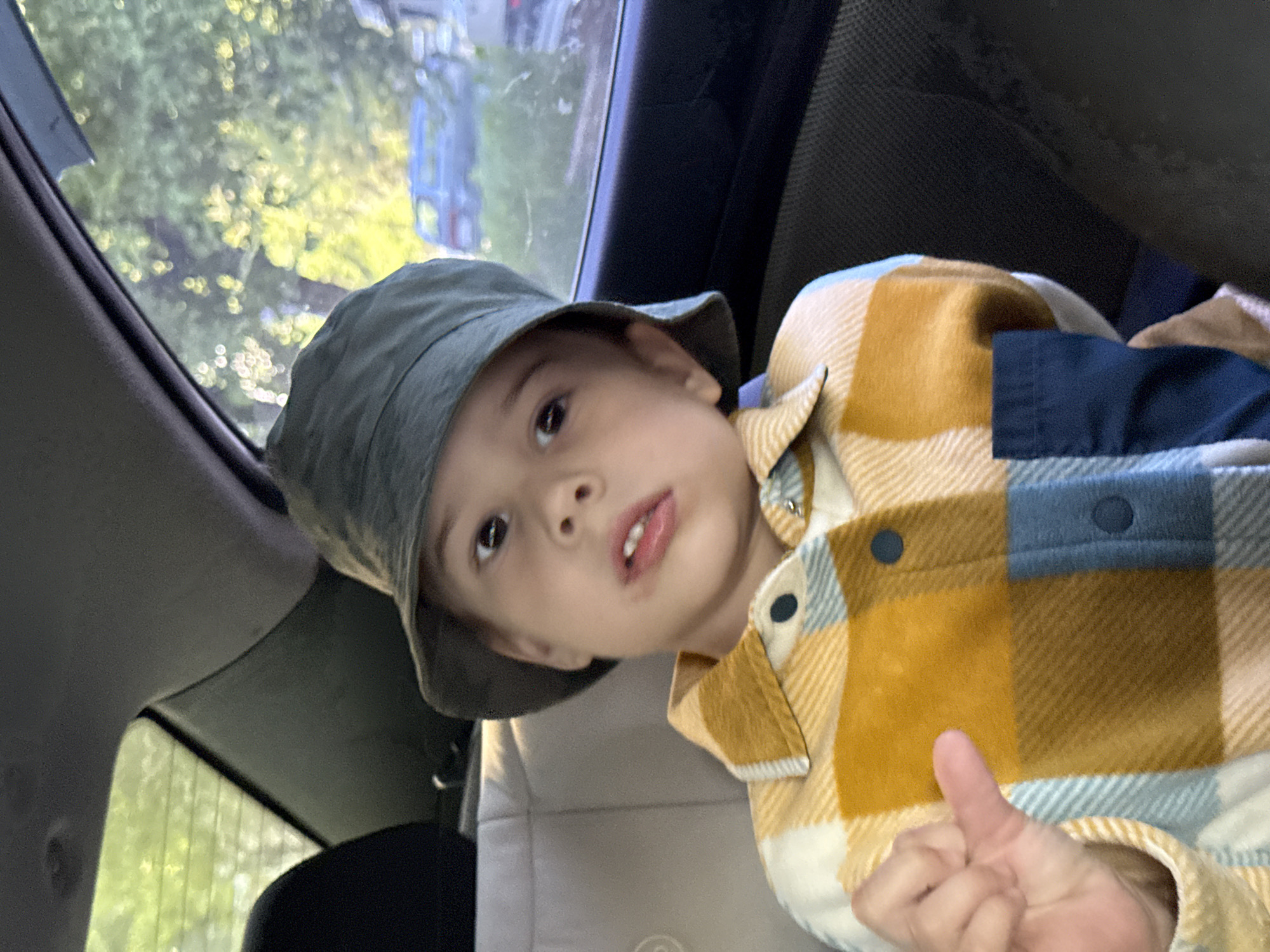 A young child wearing a grey bucket hat and a mustard yellow and blue plaid shirt is sitting in the back seat of a car. The child is giving a thumbs-up gesture. Through the car window, blurred greenery and a glimpse of a building can be seen. The location is possibly Chaikovsky, Russia.