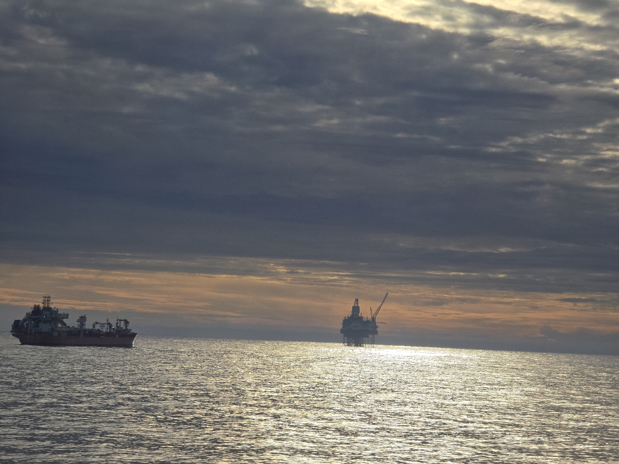 The image depicts an offshore scene under a cloudy sky, possibly at dusk or dawn. The ocean surface reflects light, creating a shimmering effect. An oil rig is situated in the distance on the right side. A ship is positioned on the left side of the image. The location is not identifiable from the provided image.