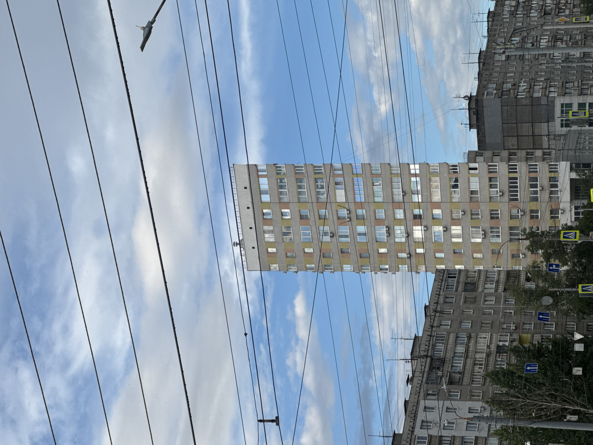 This media file depicts an outdoor scene with multiple apartment buildings under a cloudy sky. The perspective is from below, looking upwards through a series of overhead wires, possibly for trams or streetlights. The buildings are of different architectural styles and ages, with one a light-colored, multi-story structure featuring many windows, and another a darker, more imposing building with balconies and numerous windows. Trees are visible in front of the darker building, and street signs with blue arrows are present.
The time of day appears to be daytime, given the sky's brightness. The weather is partly cloudy, with patches of blue sky visible between the white clouds. The presence of apartment buildings and overhead wires suggests an urban or suburban setting. There are no people visibly active in the scene, and the focus is on the architecture and the infrastructure.
Notable details include the repetition of windows on the buildings, the contrast between the lighter and darker facades, and the geometric pattern created by the overhead wires against the sky. The image has been rotated, with the buildings appearing on the left and right sides and the sky in the center. No discernible text is visible in the image. The overall impression is that of a typical urban landscape in Russia, specifically Nizhny Novgorod, with its characteristic Soviet-era apartment blocks.