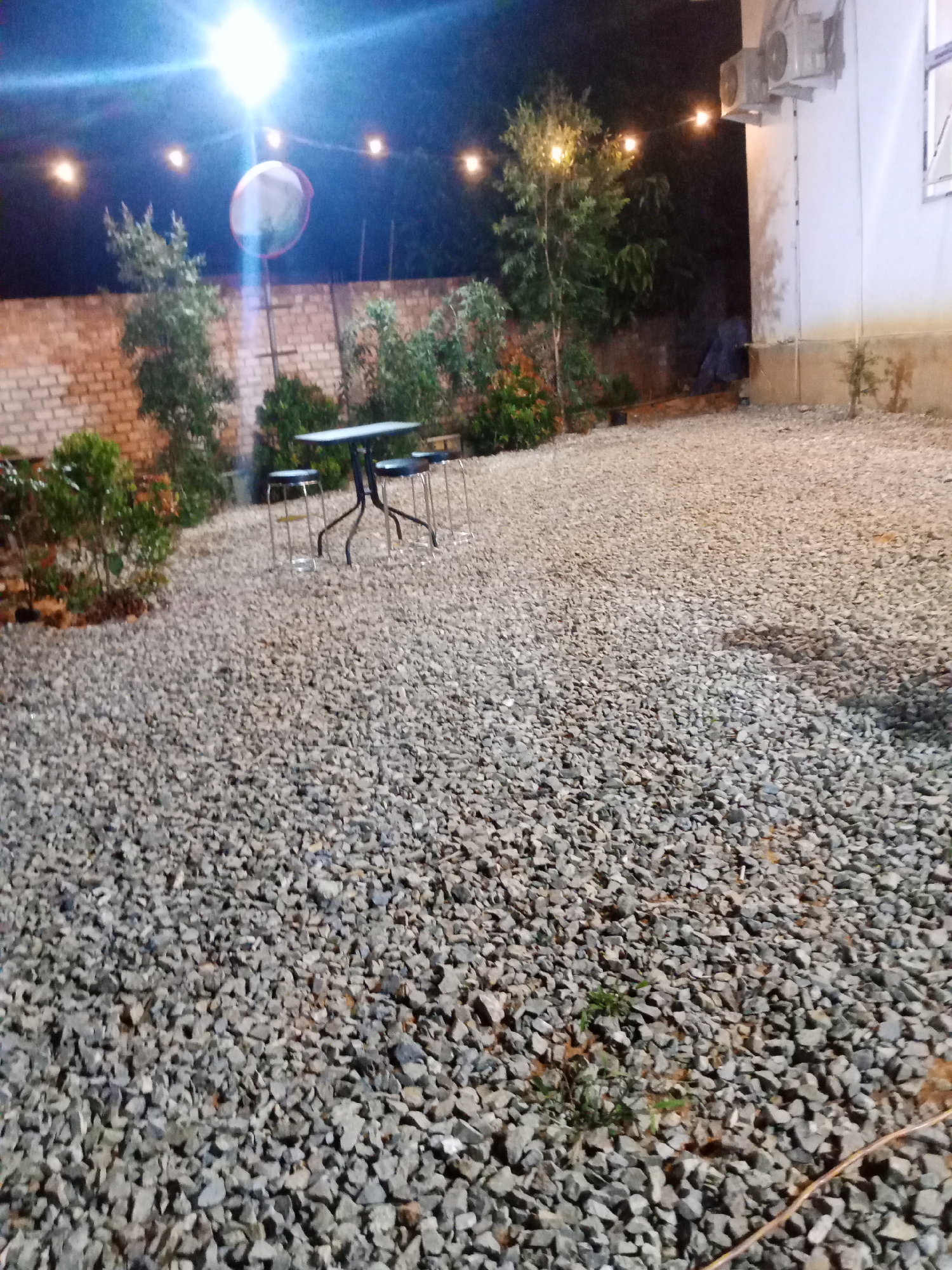 The image depicts an outdoor seating area at night, likely in Mekar Jaya, Indonesia. The ground is covered with coarse gravel. In the center of the frame, there is a dark-colored table with a dark frame, accompanied by three metal stools. To the left, a brick wall is visible, partially obscured by foliage. Above the scene, string lights are illuminated, casting a warm glow. The overall ambiance suggests a casual outdoor dining or gathering space. The presence of an air conditioning unit mounted on the exterior wall of a building on the right side of the image provides context for the building's structure.