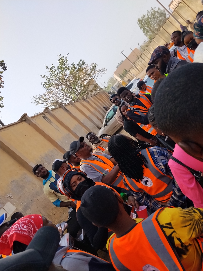 The image depicts a group of approximately 15-20 individuals gathered outdoors in Sabon Gari East, Nigeria. The scene is illuminated by natural daylight, with a pale, overcast sky visible above.
In the background on the left, a light-colored, possibly beige, concrete wall with a patterned top edge extends into the distance. Behind this wall, sparse green foliage from trees is visible. Further in the mid-ground on the right, a light-colored sedan is partially visible, parked or moving, alongside distant structures and a utility pole.
The foreground and mid-ground are populated by the group, consisting primarily of young adults. Many individuals are wearing bright orange safety vests over various casual clothing items, including t-shirts, plaid shirts, and long-sleeved tops. Some vests display a white circular logo on the back. Headwear is common, including black baseball caps, a plaid flat cap, and a red bandana worn around the neck or head. At least one female subject wears a black face mask and headphones or earmuffs. Another individual holds a red plastic cup. Several individuals are engaged in conversation or looking in various directions, some directly towards the camera's perspective. The overall posture of the group suggests a casual gathering.