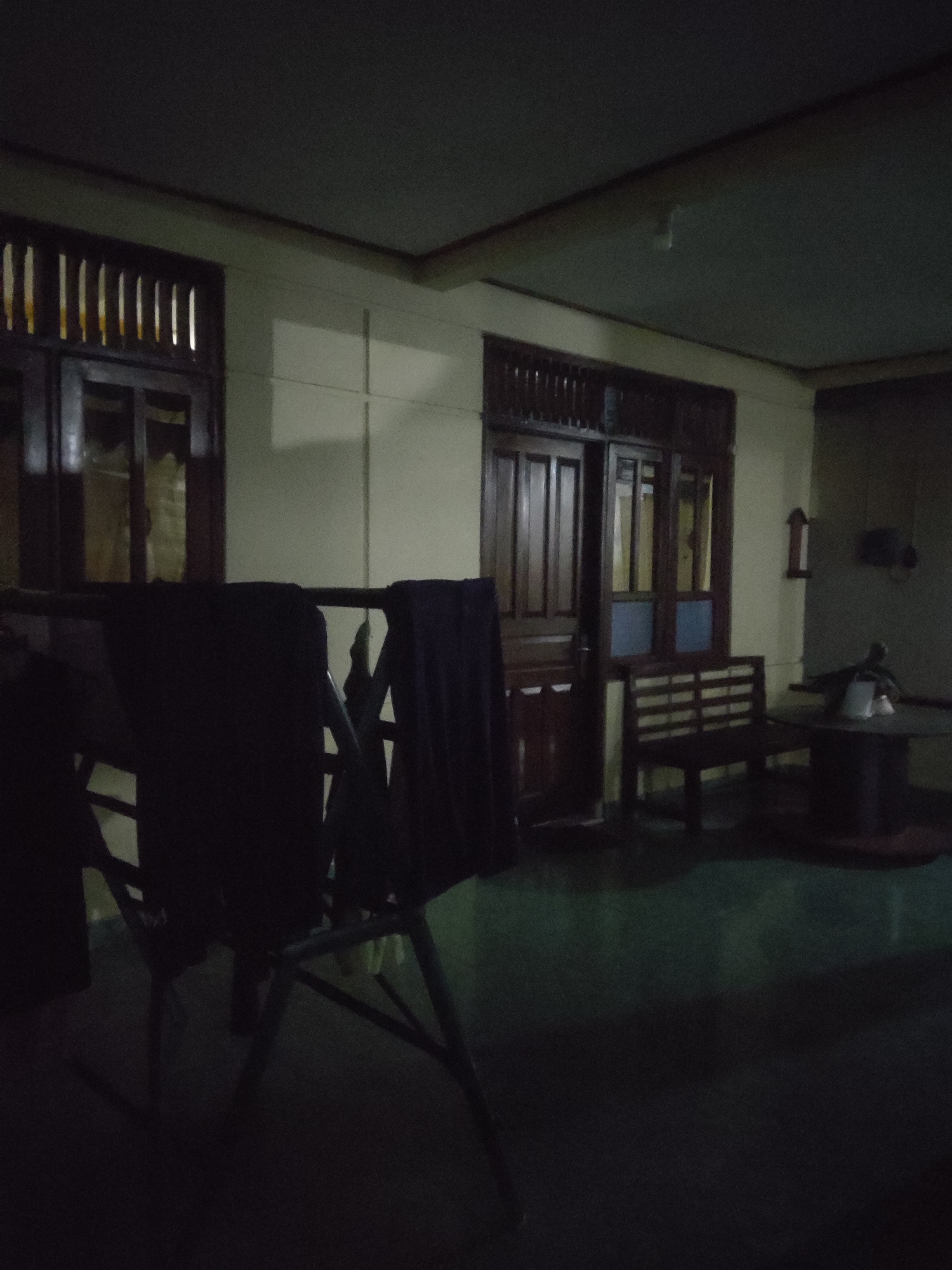 The image depicts a dimly lit, covered outdoor area, possibly a veranda or patio, in front of a building. The walls are painted a light color, and a dark ceiling is visible. To the left, there are two windows with dark wooden frames and slatted openings, suggesting they are louvred windows. In the center, a dark wooden double door with glass panes is visible. To the right of the door, a dark wooden bench with slatted seating sits on a dark floor. Further to the right, a round table with a dark base and top is present. On the table, a white object, possibly a cup or container, is visible. In the foreground, a dark metal clothes drying rack is positioned, with dark fabric draped over it. The overall impression is of a residential or common area at night, with minimal lighting. There are no people visible in the image. The scene is likely located in Trihanggo, Indonesia, given the context.