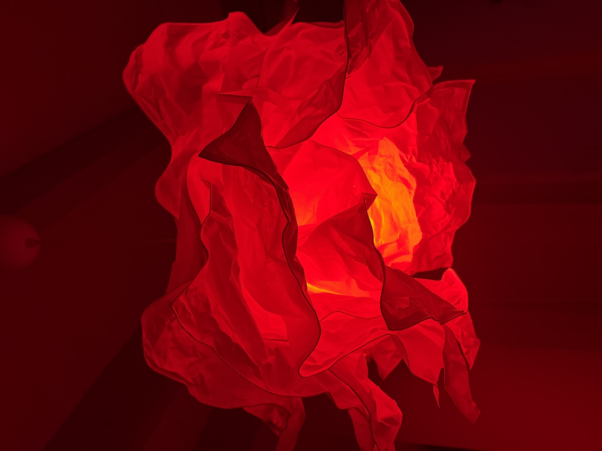 The image depicts a red, abstract, flower-like structure illuminated from within with a yellow light source. It appears to be a hanging decorative piece. The surrounding background is a darker shade of red. There is no indication of the image being taken in Chaikovsky, Russia, or any people, subjects, objects, actions, interactions, or environment that can be described beyond the illuminated red object.