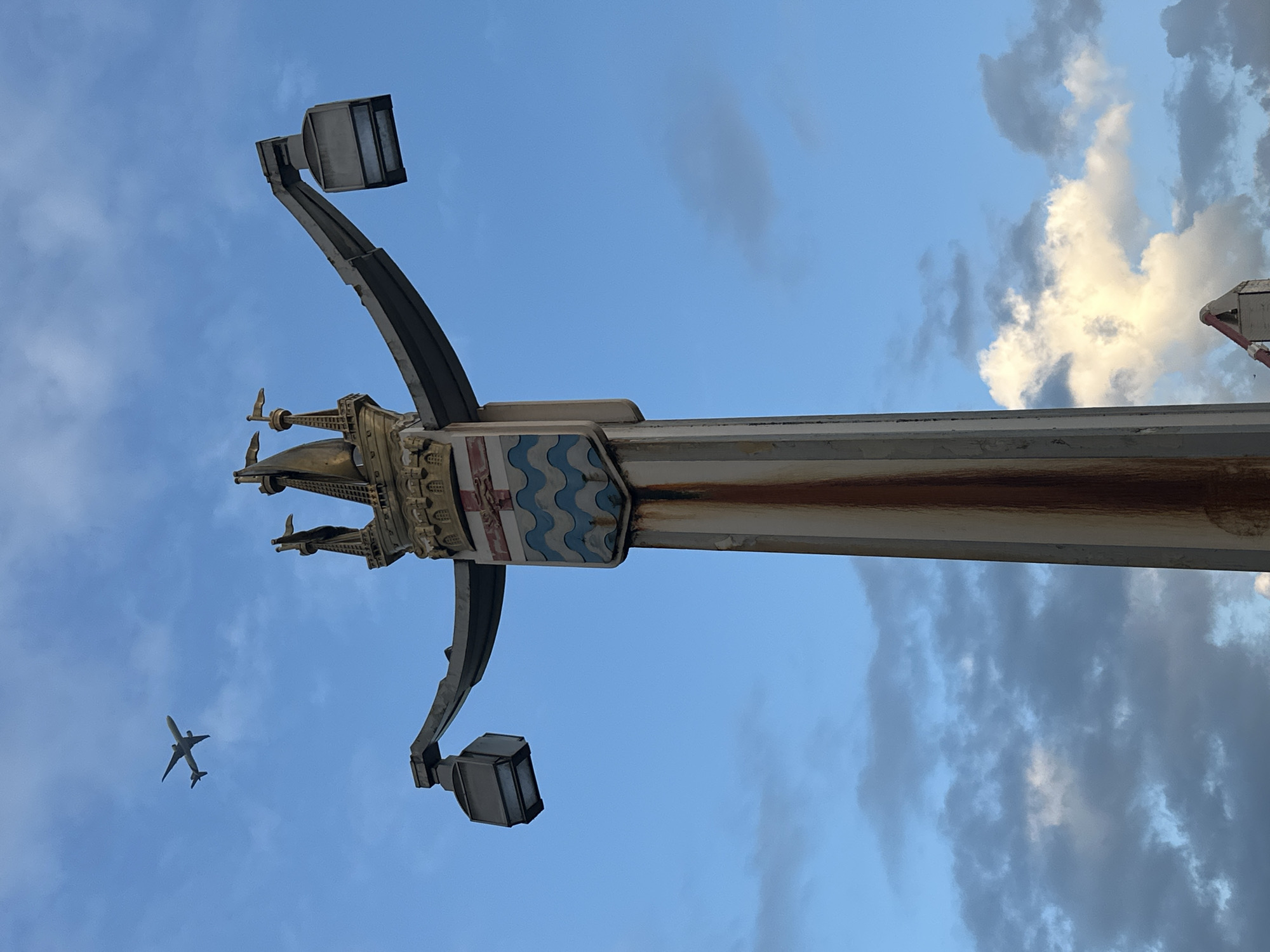 The image shows a low-angle view of a street lamp in London, United Kingdom, set against a partly cloudy sky. The lamp post is ornate, featuring a shield with blue and white wavy lines. Two lamp fixtures extend from either side. An airplane is visible in the lower left corner of the frame.