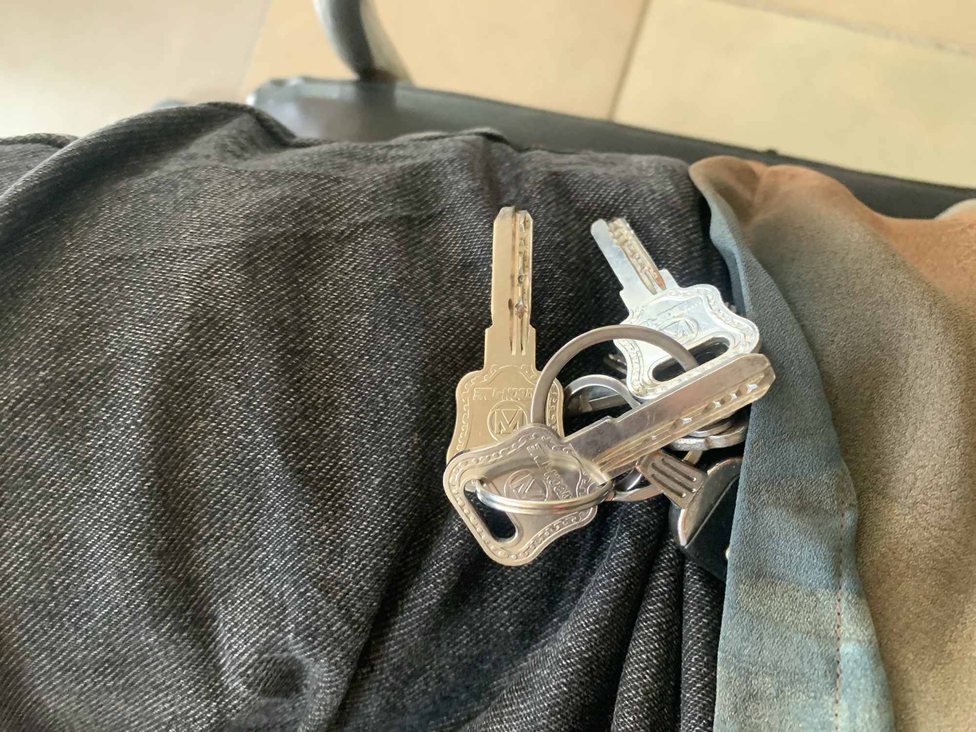 A close-up, eye-level shot shows a bunch of silver keys held together by a keyring, resting on dark gray denim material. The keys have intricate detailing and appear to be for multiple locks. The denim is creased and folded, suggesting it is part of a garment like pants. Part of a light brown material, possibly leather or suede, is visible underneath the denim. The background is out of focus but suggests an indoor setting with a light-colored wall and a dark object, possibly a chair. The lighting is even, with no strong shadows.
