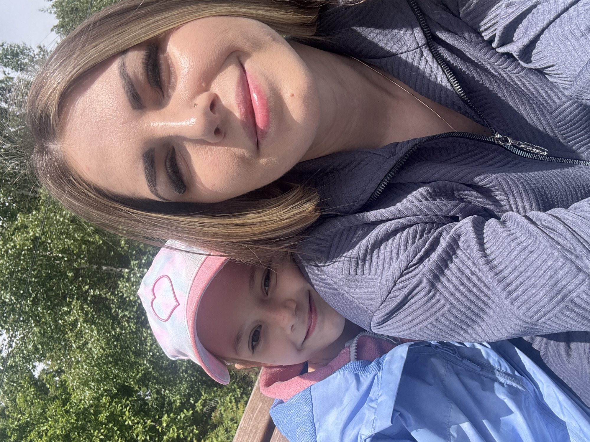 A woman and a young girl are posing for a close-up, angled selfie. The woman has light brown hair with blonde highlights, fair skin, closed eyes, and pink lips. She is wearing a gray, textured zip-up jacket. A thin necklace is visible on her neck. The young girl is positioned below the woman, wearing a pink and white baseball cap with a heart design, and a light blue jacket with a pink interior hood. The background shows green trees. The image suggests an outdoor setting, possibly in Shevlyakovo, Russia.