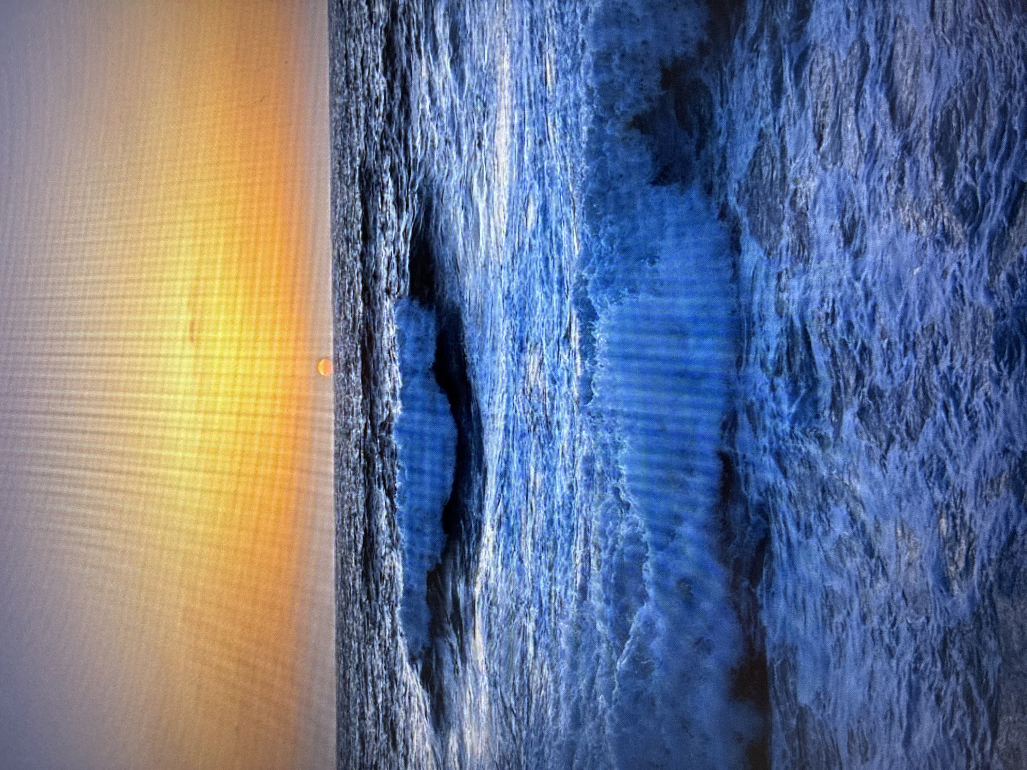 The image depicts a scene where a body of water, potentially the ocean, meets the shore. The left side of the image showcases a soft, orange glow, suggestive of either sunrise or sunset. The water is turbulent with visible waves crashing and churning. No people, objects, actions, or interactions are clearly identifiable in the image. There is no evidence in the image to place it in Bat Yam, Israel.