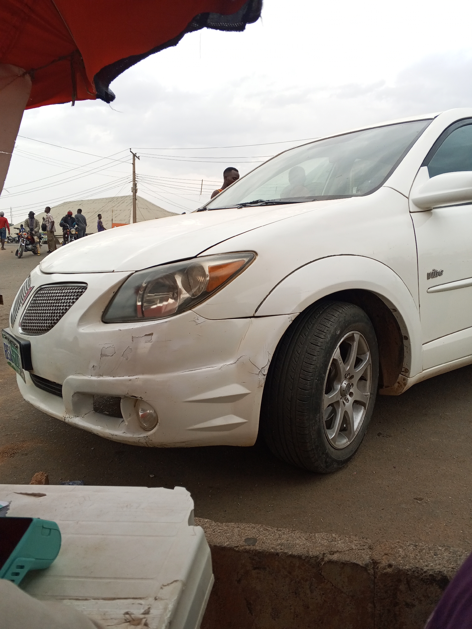 The image depicts a white Pontiac Vibe parked on a paved road in Gwong, Nigeria. The car's front bumper has visible paint damage. In the background, several people are standing near motorcycles on a dirt area. Above them is a grey sky. A portion of a red and black awning is visible in the upper left corner. A white plastic container and a teal object (possibly a phone) are in the foreground.