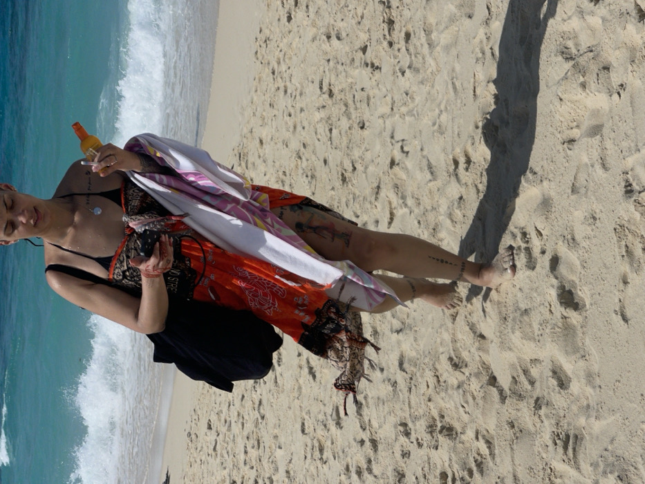 A solitary individual is observed walking barefoot on a sandy beach in the city of Zanzibar, Tanzania. The subject, an adult female with visible tattoos on her arms and legs, wears a black bikini top beneath an orange and black patterned sarong. A black shoulder bag is carried on her left shoulder. In her left hand, she holds a dark-colored smartphone. Her right hand grasps an orange bottle, likely sunscreen, and a small white object, while a white and multi-colored striped towel is draped over her right forearm. The individual casts a long shadow extending to her right on the sand. The environment consists of light-colored sand in the foreground and midground, transitioning to turquoise ocean water with white breaking waves in the background.