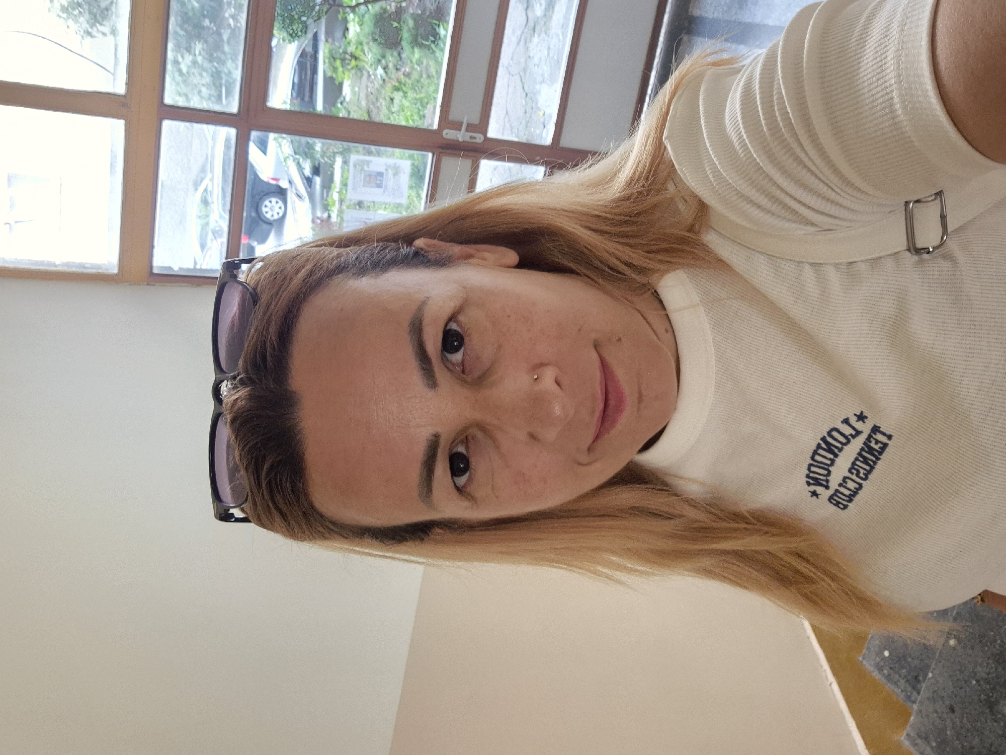 A medium shot captures a woman with blonde hair and glasses perched on her head, facing forward. She wears a white, ribbed t-shirt with a blue embroidered logo that reads "LONDON TENNIS CLUB". Her left shoulder is visible, adorned with a metal buckle detail on a strap. The background features a large window with wooden frames, revealing a glimpse of cars and greenery outside. The lighting is natural, coming from the window. The scene appears to be indoors, possibly in a hallway or near a staircase, indicated by the textured wall and a sliver of stairs in the lower-left corner.