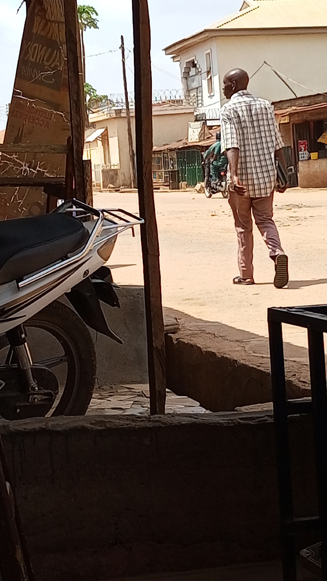 The image shows a street scene in Sabon Tasha, Nigeria. In the foreground, part of a white motorcycle and a weathered, wooden structure with faded text are visible. The middle ground features a man with a bald head, wearing a checkered shirt and reddish-brown pants, walking away from the camera. He is holding something dark in his left hand. In the background, buildings with corrugated metal roofs and barbed wire are visible, as well as another motorcycle with two occupants. The street is unpaved and dusty.