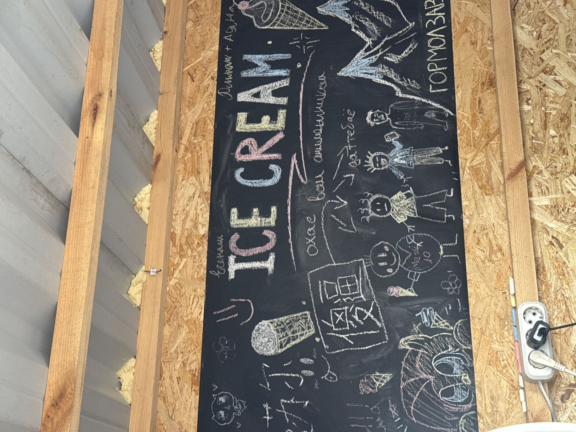 The image is a close-up, slightly angled shot of a blackboard with chalk drawings and text. The blackboard is mounted on a wooden wall, and above it, exposed wooden beams and insulation are visible, suggesting an outdoor or unfinished interior space. The blackboard itself is covered with various drawings, including ice cream cones, cartoon characters, and what appear to be mountains. The main text on the blackboard reads "ICE CREAM" in large, colorful letters. Below "ICE CREAM" is some smaller, handwritten text in what appears to be Cyrillic script. To the right of the "ICE CREAM" text, there is a drawing of an ice cream cone, and further to the right, a mountain range is depicted. The bottom section of the image shows a power strip with two plugs inserted, mounted on a wooden surface. There are also some chalk sticks in various colors lying on the wooden surface in front of the power strip. The overall impression is of a casual, possibly temporary setup for an ice cream vendor. The location is Kokshetau, Kazakhstan.