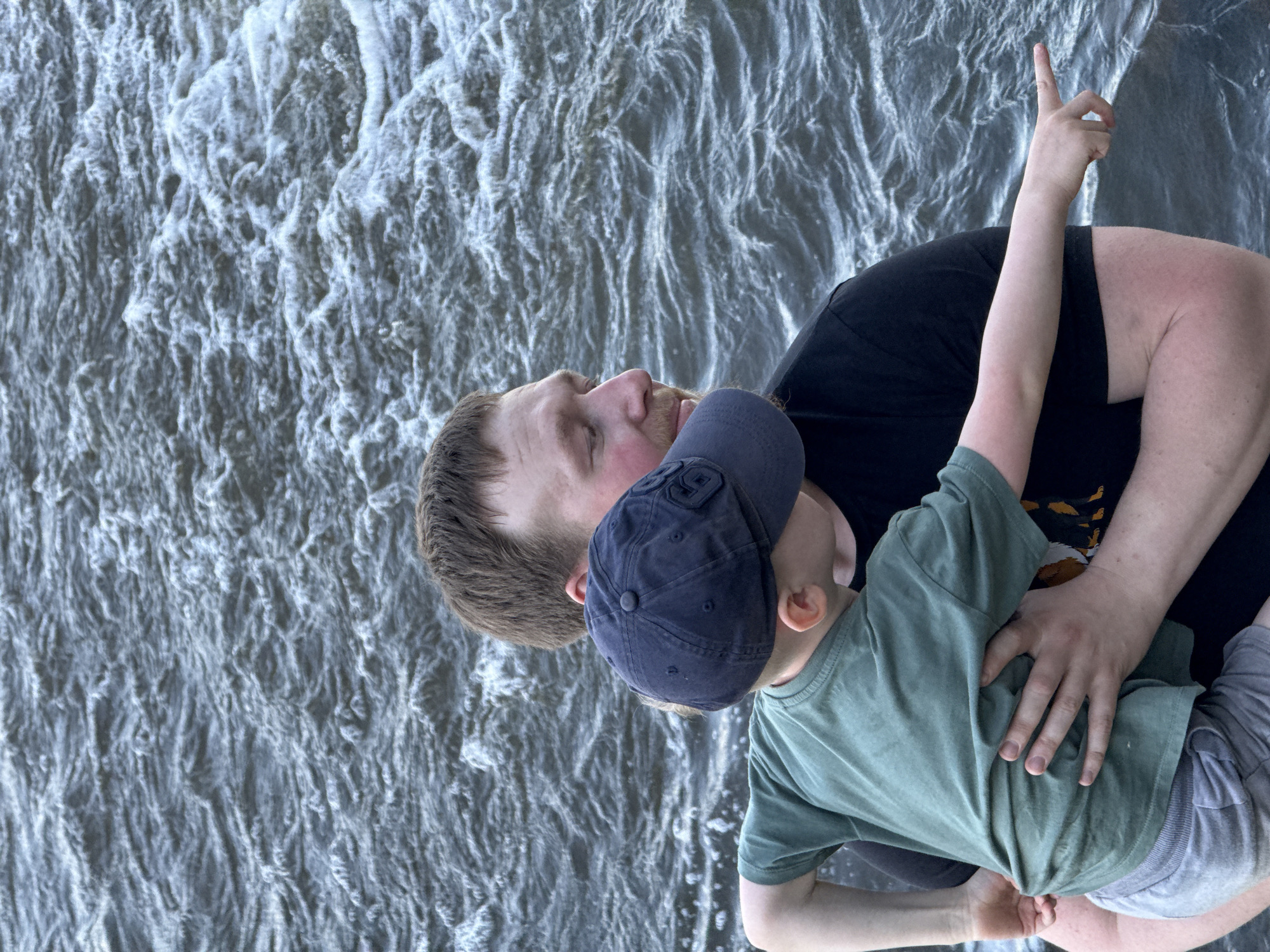 A man and a young child are standing in front of a body of water, possibly a fast-moving river or stream. The man is wearing a black t-shirt, and the child is wearing a green t-shirt and a dark-colored baseball cap. The child has one arm raised, pointing upwards, and the man is looking in the same direction. The water behind them is turbulent. The location is Novyi, Russia.