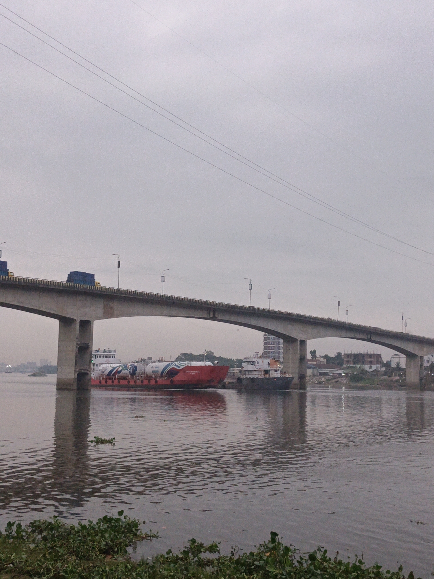 The image depicts a concrete bridge spanning a wide river in Dhaka, Bangladesh, under an overcast sky. The bridge carries vehicular traffic. A red and white tanker ship navigates the river beneath the bridge, with a smaller boat nearby. The river's surface is rippled, reflecting the light. Aquatic plants grow along the riverbank in the foreground. Buildings are visible in the background on the riverbank. Electrical wires are visible above the bridge.