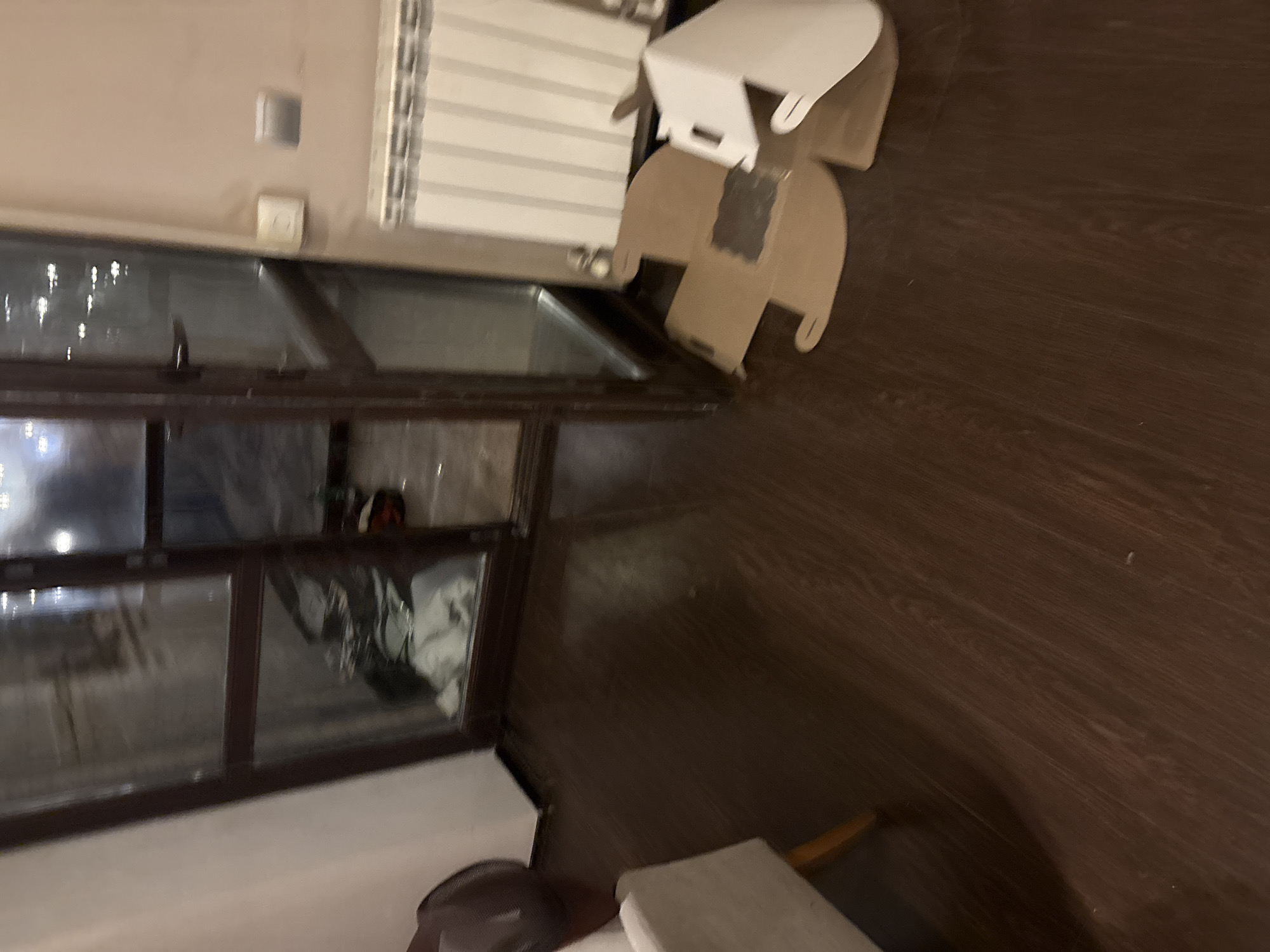 The image depicts an interior scene, likely a room in a residential building. The floor is made of dark wood laminate. To the left, a portion of a light-colored armchair is visible. Dominating the center and right of the image is a dark wooden door with glass panels. Through the glass, a dimly lit interior space, possibly a balcony or another room, is visible, containing some folded white fabric and dark items.
In the foreground, on the right side of the door, are several pieces of flattened cardboard boxes and a white packaging material, suggesting recent unpacking or disposal. A white radiator is mounted on the wall above these boxes. To the right of the door, on the wall, there are two light switches or electrical outlets. The overall lighting is dim and artificial. The scene appears to be indoors, possibly in a living area or hallway, in a city like Orekhovka, Russia, though no specific landmarks confirm this location. There are no people visible in the image.