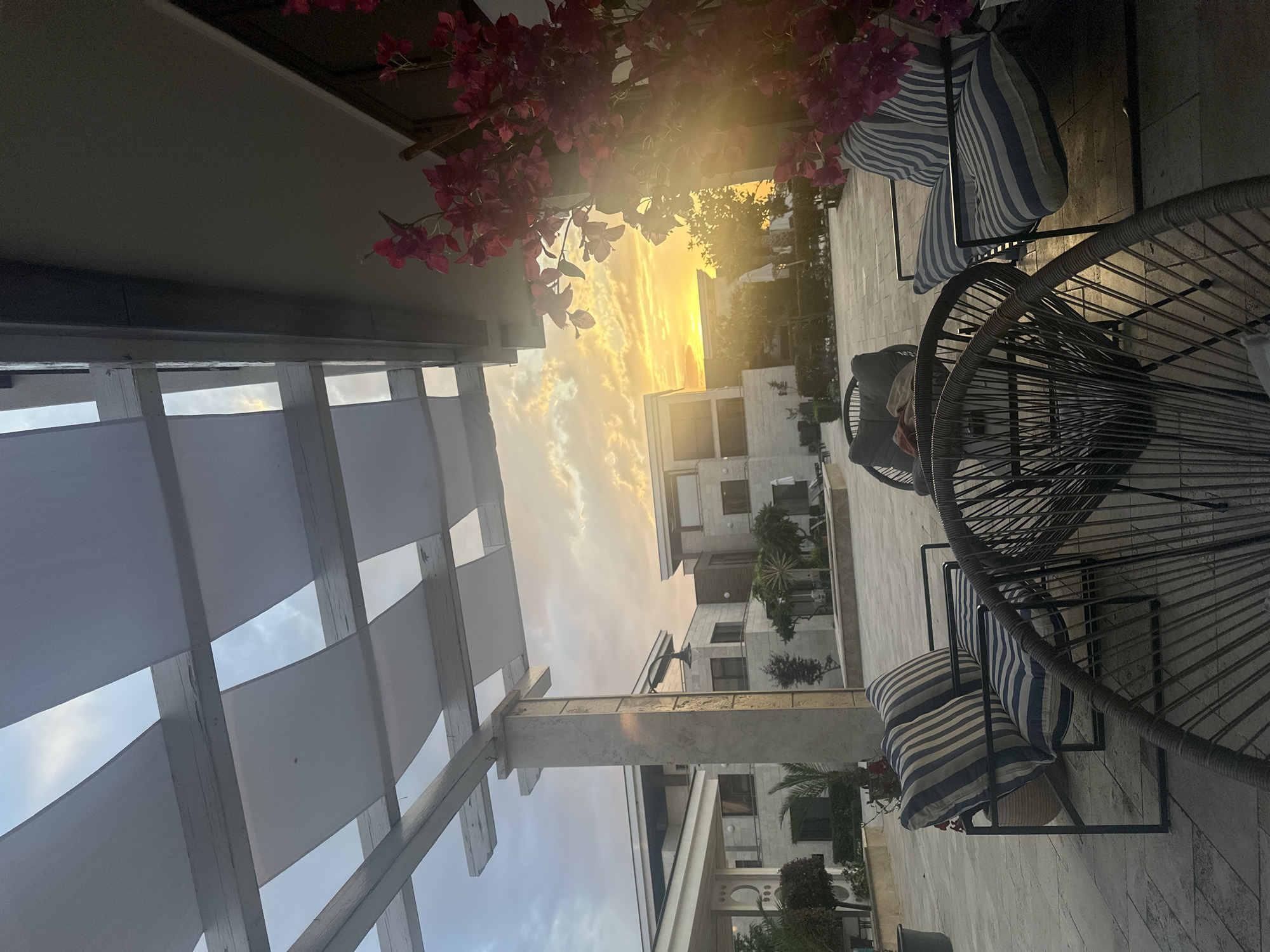 This media file captures an outdoor patio scene at sunset in Sozopol, Bulgaria. The focal point is a seating area with two distinctive wireframe chairs, each adorned with blue and white striped cushions. One chair appears empty, while the other has a few items on it, including what looks like a basket and possibly a towel. Adjacent to the chairs, a pergola structure with white fabric panels overhead suggests a shaded outdoor space.
The background reveals a modern architectural setting, with buildings featuring stone facades and balconies. Lush greenery, including palm trees and smaller bushes, is interspersed around the buildings, hinting at a warm climate. The sky is dominated by a dramatic sunset, with vibrant oranges and yellows illuminating the clouds, casting a warm glow over the entire scene. A cluster of vibrant pink bougainvillea flowers hangs from a branch in the upper left portion of the frame, adding a splash of color against the softer tones of the sky and architecture.
The overall atmosphere is one of tranquility and relaxation, evoked by the peaceful setting and the beautiful sunset. The time of day is clearly evening, as indicated by the sunset. The weather appears to be pleasant, given the outdoor seating and the clear visibility. No people are visible in the frame, and there is no discernible text. The composition, with its tilted perspective, adds a dynamic element to the otherwise serene scene.