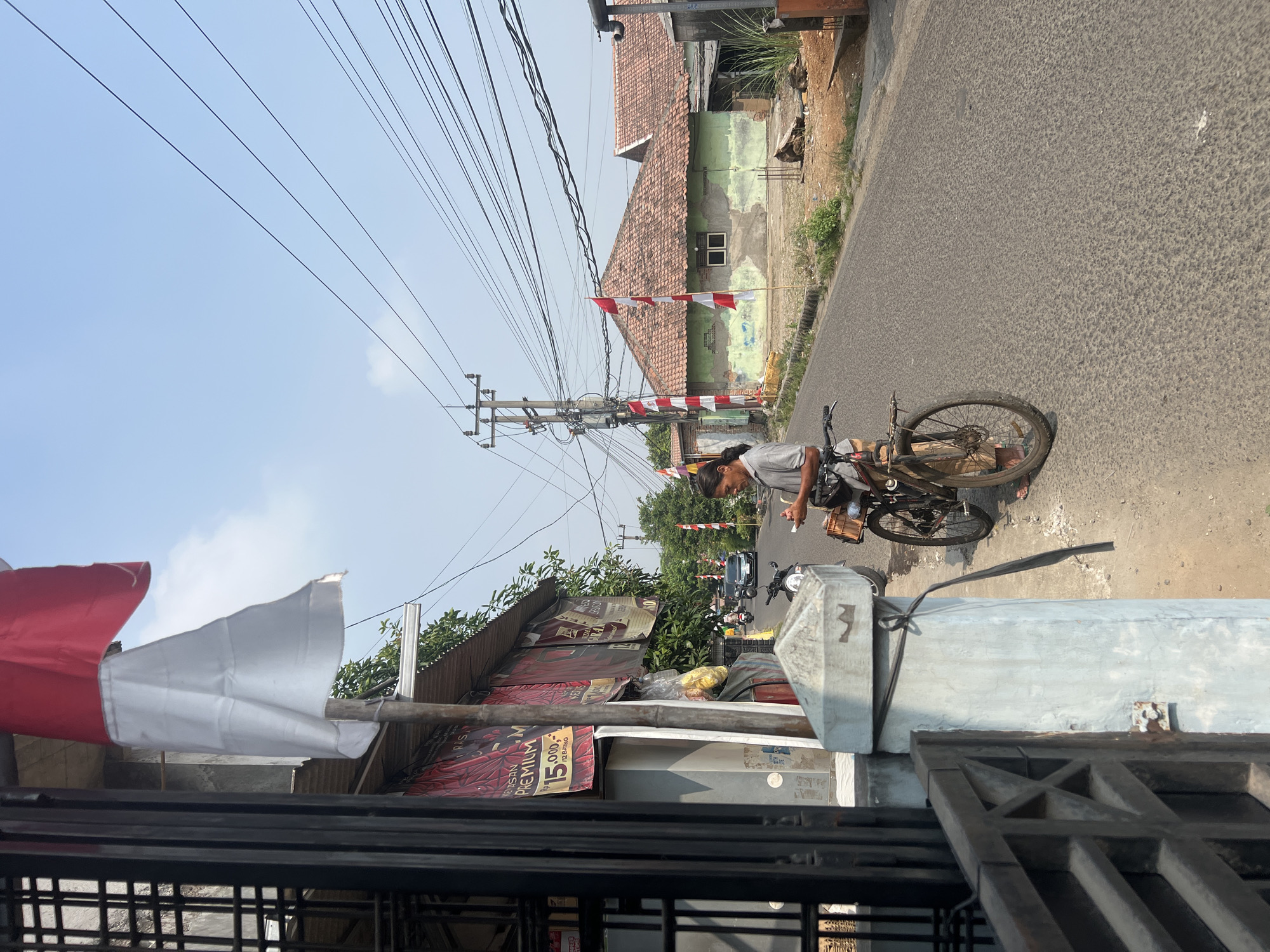 This is a street-level view of a residential area in Klapanunggal, Indonesia. In the foreground, a man stands next to a bicycle, looking at his phone. The bicycle is a dark color with a basket on the back. To the left, a metal gate and a pillar with a wire wrapped around it are visible. Behind the pillar is a building with red and white banners displayed. Across the street, two houses are visible. The house on the left is green with a red tiled roof, and a small Indonesian flag is planted in front of it. The house on the right also has a red tiled roof. The street is paved and appears to be in moderate condition, with some debris on the side. Overhead, a complex network of power lines crisscrosses the sky. The sky is clear and blue. In the background, a few motorcycles and a car are driving on the street. The overall impression is of a typical Indonesian neighborhood on a sunny day.