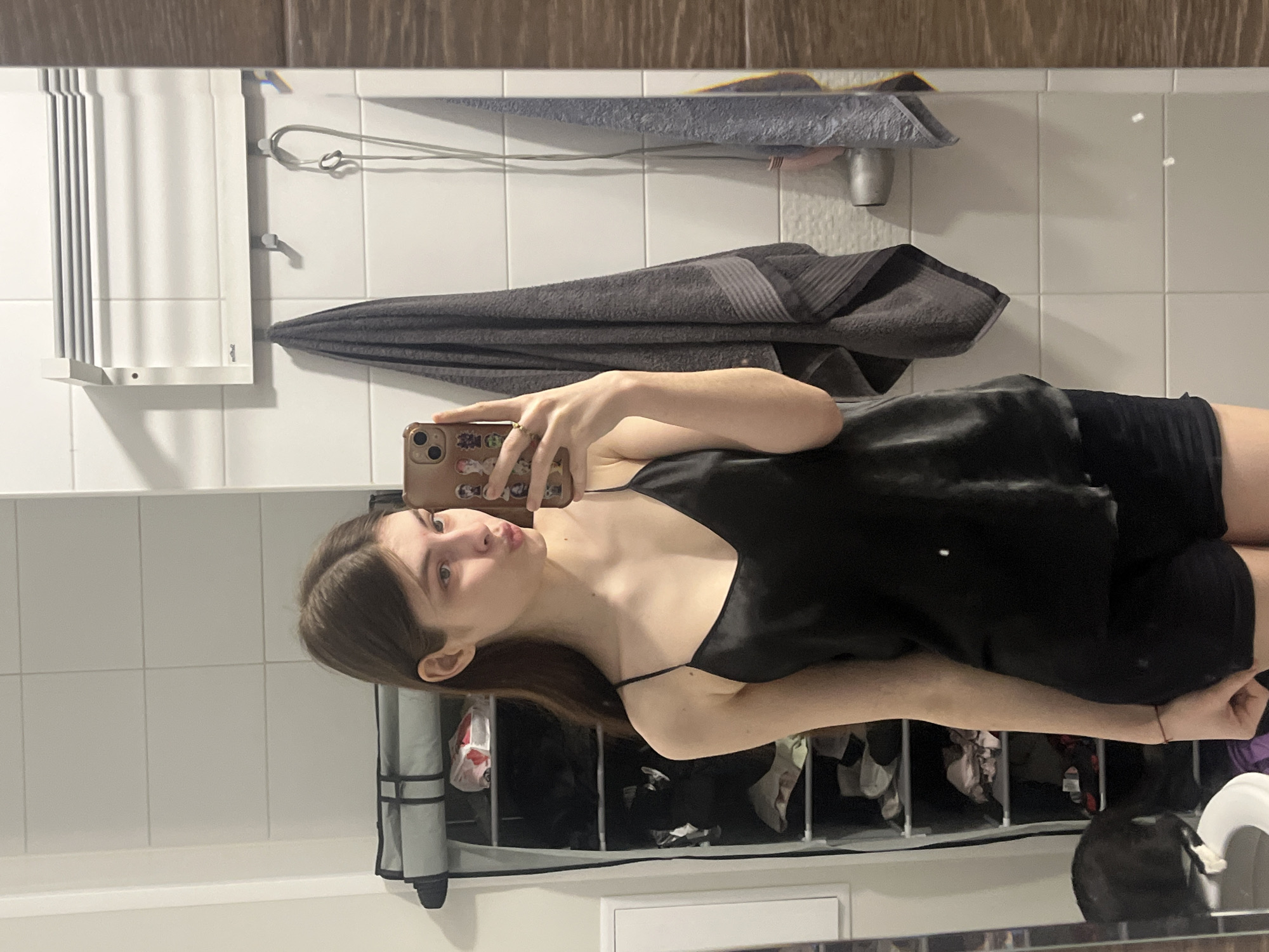 A woman is taking a selfie in a bathroom mirror. She has brown hair and is wearing a black camisole top and black shorts. The background reveals white square tiles on the wall, a white towel warmer, a gray towel, and a rack with clothes. A black cat is visible in the lower right corner. There is no indication that this image was taken in Saint Petersburg, Russia.