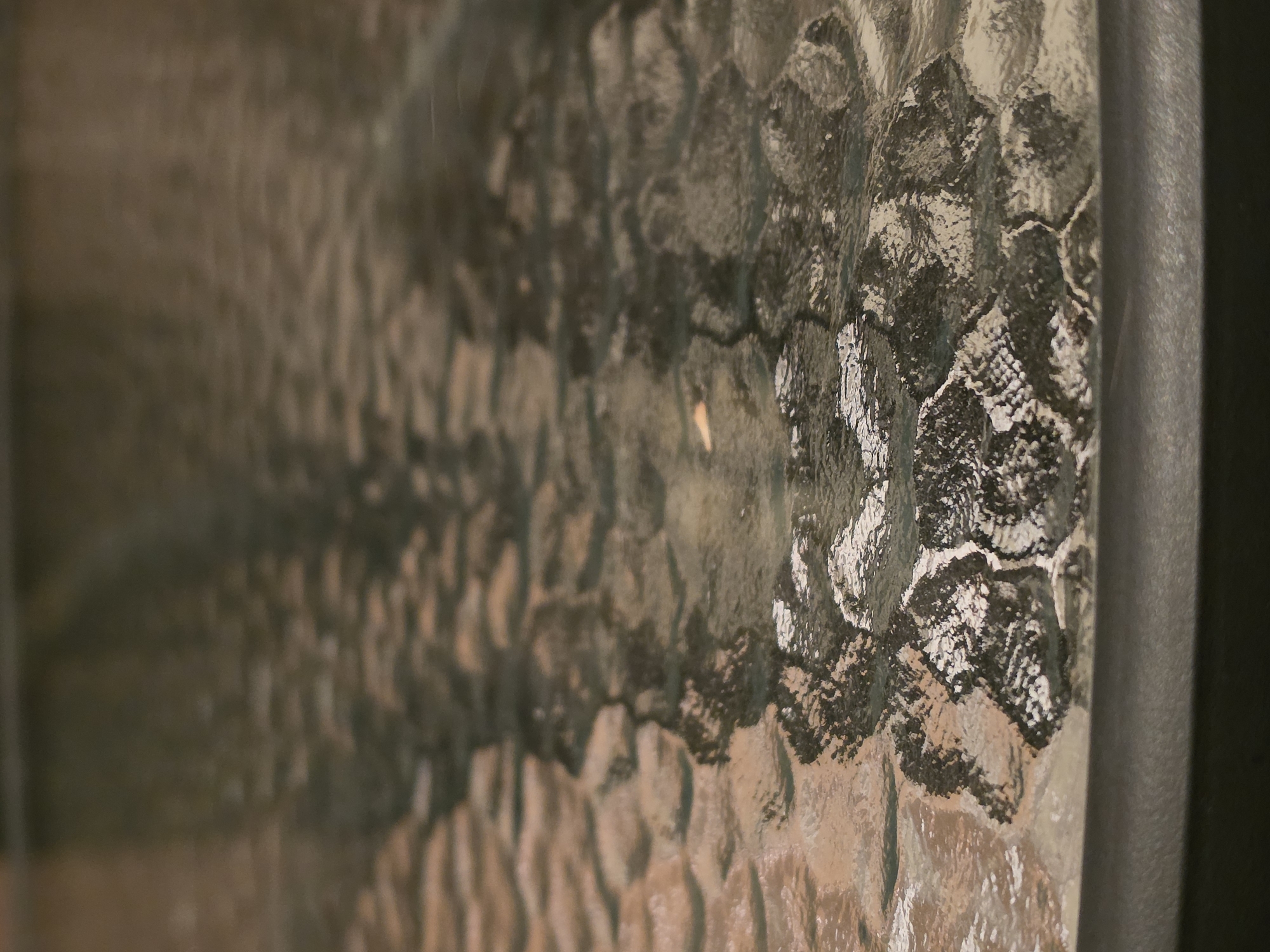 A close-up, slightly elevated shot focuses on the textured, patterned surface of a dark, possibly glass, table. The surface is mottled with dark and light areas that suggest a patterned texture or reflection. The foreground shows a rounded, dark edge of the table. The background is blurred, indicating a shallow depth of field, and consists of indistinct darker shapes and lighter, streaky patterns. The scene appears to be indoors, in a location in the city of Nagorny, Russia, though no specific landmarks or identifying features of the city are visible in the image.