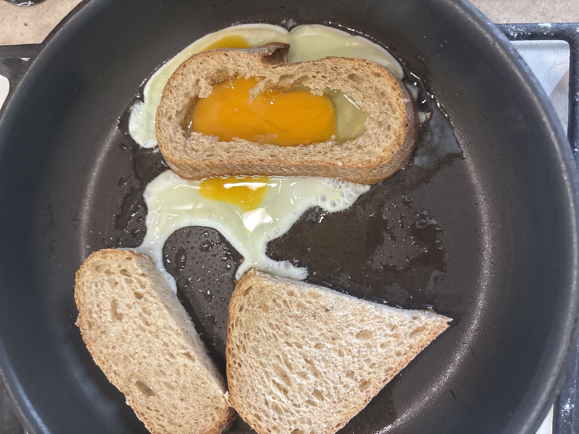 An overhead shot shows two slices of bread and one slice of bread with a hole cut out of it, all in various stages of cooking in a black frying pan. The slice with the hole has a raw egg yolk in its center, surrounded by its white. The other two slices are partially submerged in the egg white. The scene is a kitchen, likely in Bila Tserkva, Ukraine, where someone is preparing a breakfast dish.