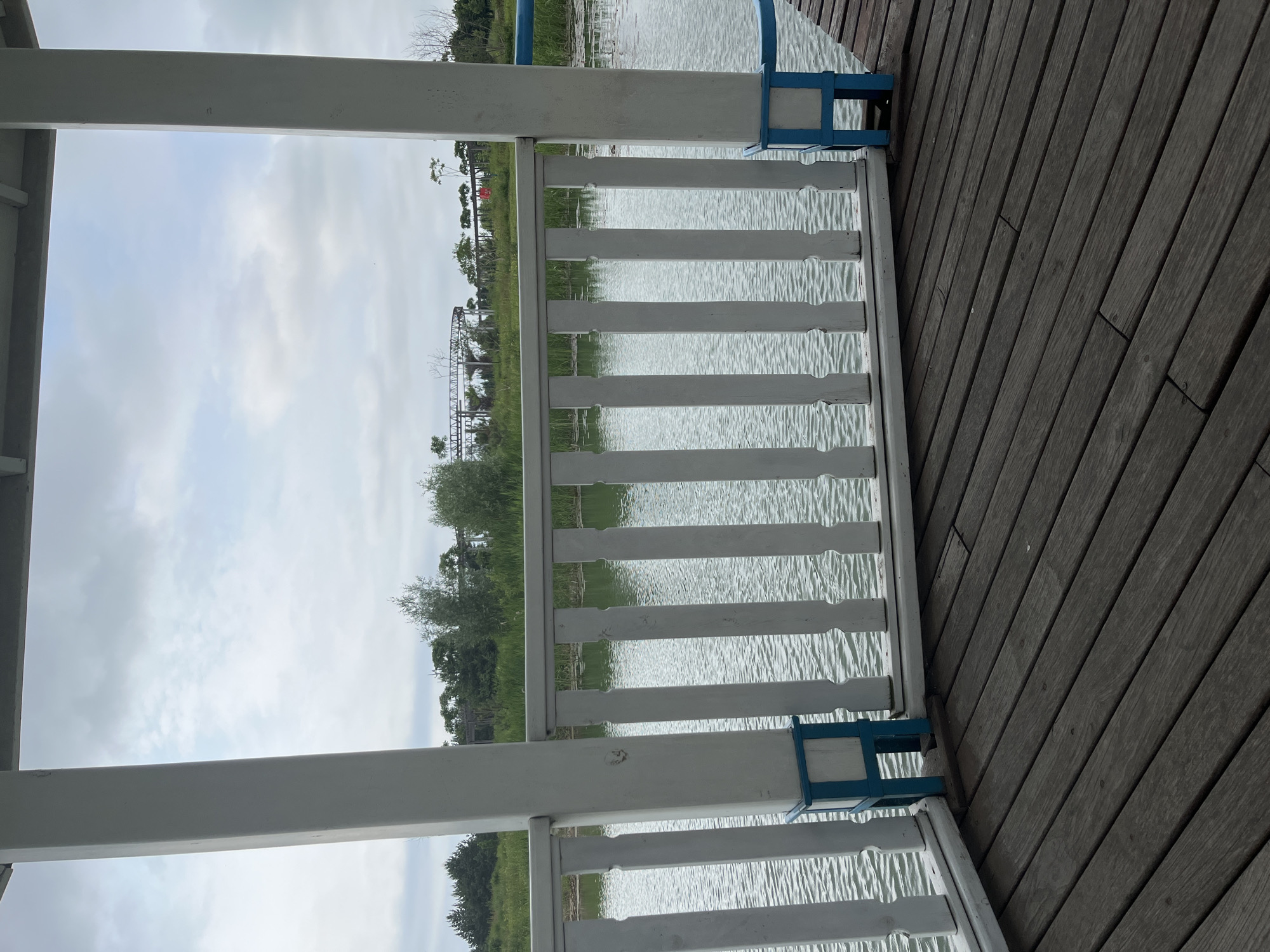 The image captures a view from a wooden deck, looking out towards a body of water. A white wooden railing with blue accents separates the deck from the water. Beyond the railing, the calm water reflects the overcast sky. In the distance, a line of green trees and foliage marks the horizon. Some man-made structures, possibly part of a park or amusement area, are visible amongst the trees, including a circular track and some elevated walkways. The scene is situated in Severynivka, Ukraine, as indicated by the provided context. The overall impression is one of a peaceful, outdoor setting, likely a recreational area.