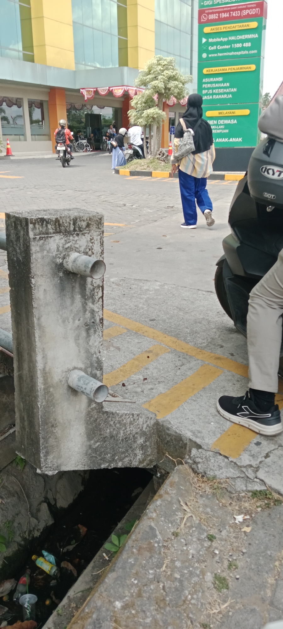 The image depicts an outdoor scene in front of a modern building, likely a hospital, in Maguwoharjo, Indonesia. In the foreground, a concrete structure with two metal pipes protruding from it is prominently featured. Below this structure is a dark opening of a drainage ditch filled with debris, including plastic bottles and other trash. The ground is paved with gray asphalt, marked with yellow lines.
In the middle ground, several people are visible. A person on a motorcycle is moving away from the camera. Another person wearing a hijab and a striped shirt, along with blue pants, is walking towards the building. A person in a wheelchair is near the entrance of the building, and other individuals are also present in front of the entrance, some near motorcycles. A large green sign with white and yellow text is visible on the right side of the frame, providing information about services and contact details for "HALOHERMINA" and "HERMINAHOSPITALS.COM".
The background shows the facade of the building, characterized by large glass windows and yellow accents. Some trees and decorative plants are also present near the entrance. The sky is overcast, suggesting a cloudy day. The overall scene captures a moment in a public space, highlighting urban infrastructure and human activity.