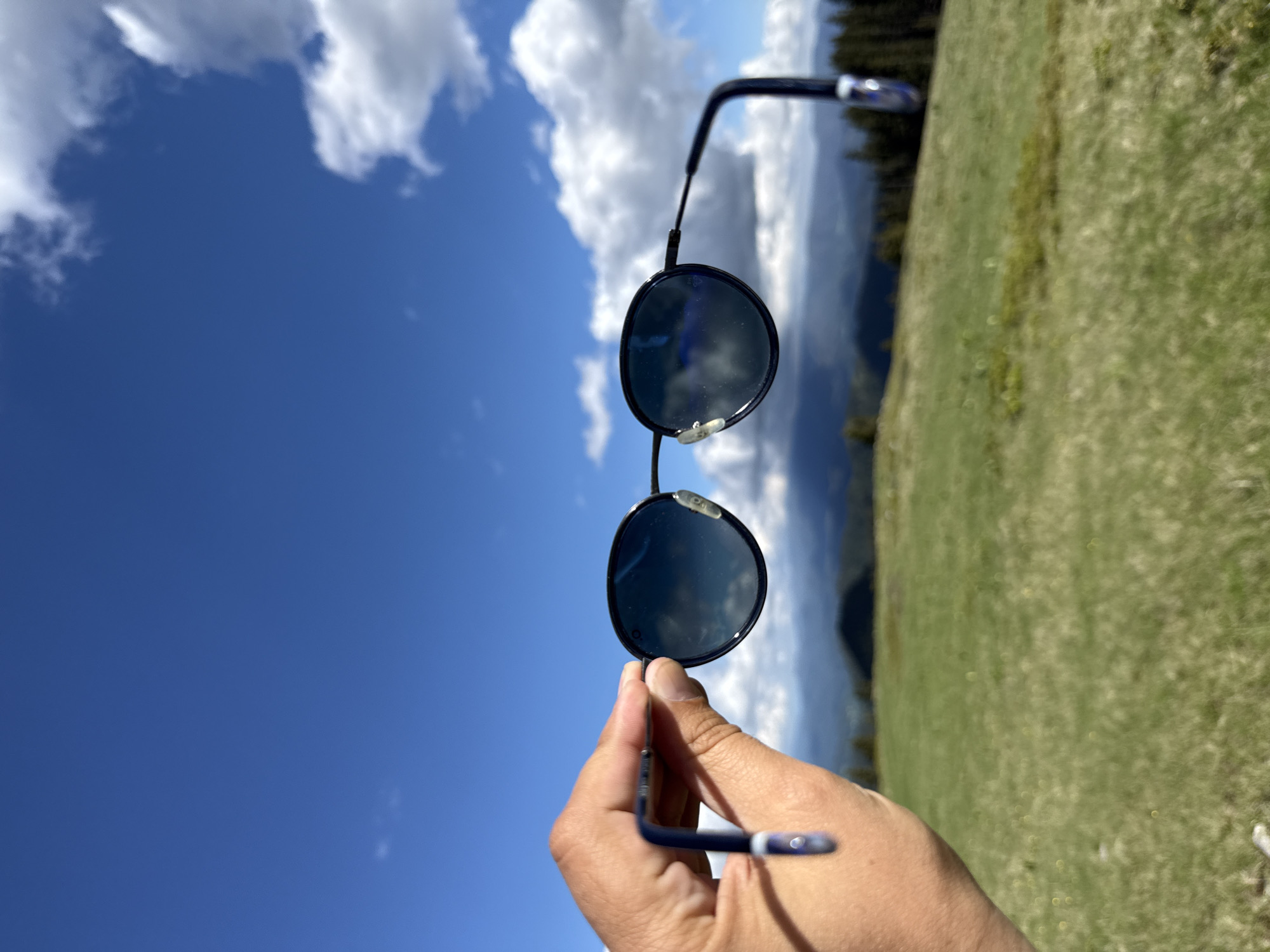 A hand holds a pair of dark-rimmed sunglasses up to the sky, obscuring the view of a mountainous landscape below. The sunglasses have round, dark lenses that reflect the blue sky and white clouds. The hand is in focus, with visible skin texture and fingernails. The background is a panorama of rolling green hills and distant, dark mountains, dotted with evergreen trees. The sky is a vibrant blue, with scattered fluffy white clouds. The image appears to have been taken outdoors in a natural setting, possibly in Gela, Bulgaria, a village known for its scenic mountain views.