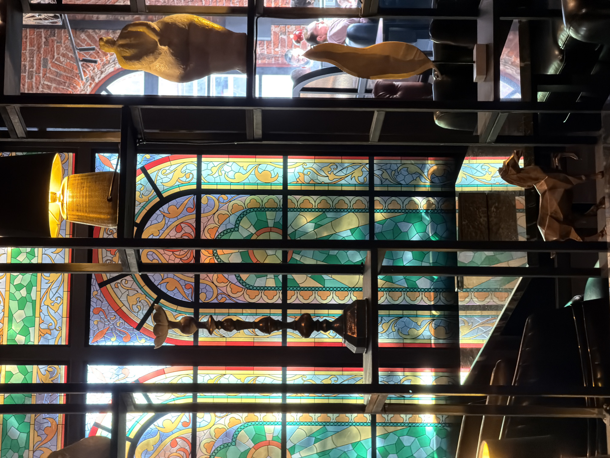 The image shows a view of a complex interior space, likely a restaurant or bar in Moscow, Russia, judging by the architectural details. The focus is on a stained-glass ceiling or window arrangement with geometric and floral patterns in shades of green, blue, yellow, and red. A black metal framework supports the glass structure.
Hanging or mounted within the framework are various decorative objects, including a yellow-toned lamp with a mesh lampshade, a long decorative metal element that resembles a stylized candelabra or architectural flourish, a paper-like sculpture of an animal (possibly a wolf or dog), and two plaster casts - one shaped like a hand and the other a curved form resembling a leaf or stylized shell.
Above this arrangement, in another level, one can see blurred figures of people seated at tables, suggesting a mezzanine or upper floor area. The overall environment gives a sense of eclectic decor and artistic design.