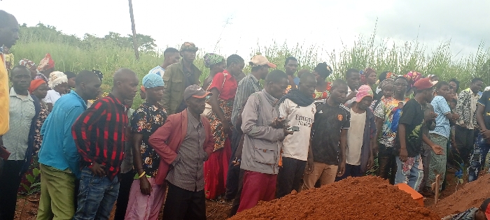 A crowd of people stands alongside a mound of reddish-brown soil, presumably a freshly dug grave. Many wear casual clothing and hats. Some are looking down at the mound. The landscape behind the group is grassy, with trees in the distance under a cloudy sky. The scene suggests a burial or memorial service.