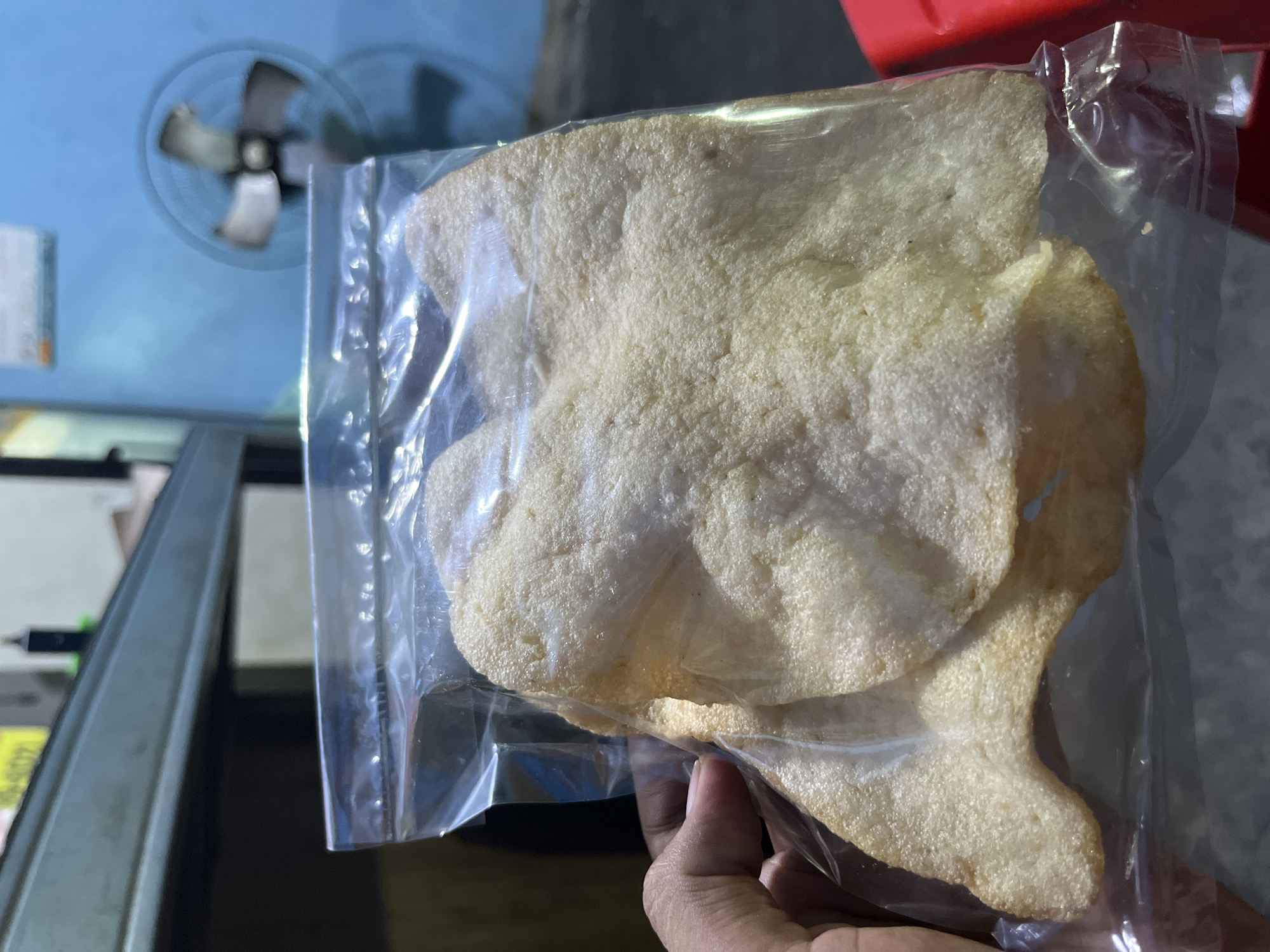 A hand is holding a clear plastic bag filled with light beige colored crackers. The crackers have an irregular, puffed, and slightly bubbly texture. The bag is sealed with a plastic strip. The background is out of focus and shows a blue wall with a black fan mounted on it, and a dark colored shelf. A red object is partially visible on the right side of the image. The scene appears to be indoors. The image was taken in Central Jakarta, Indonesia.