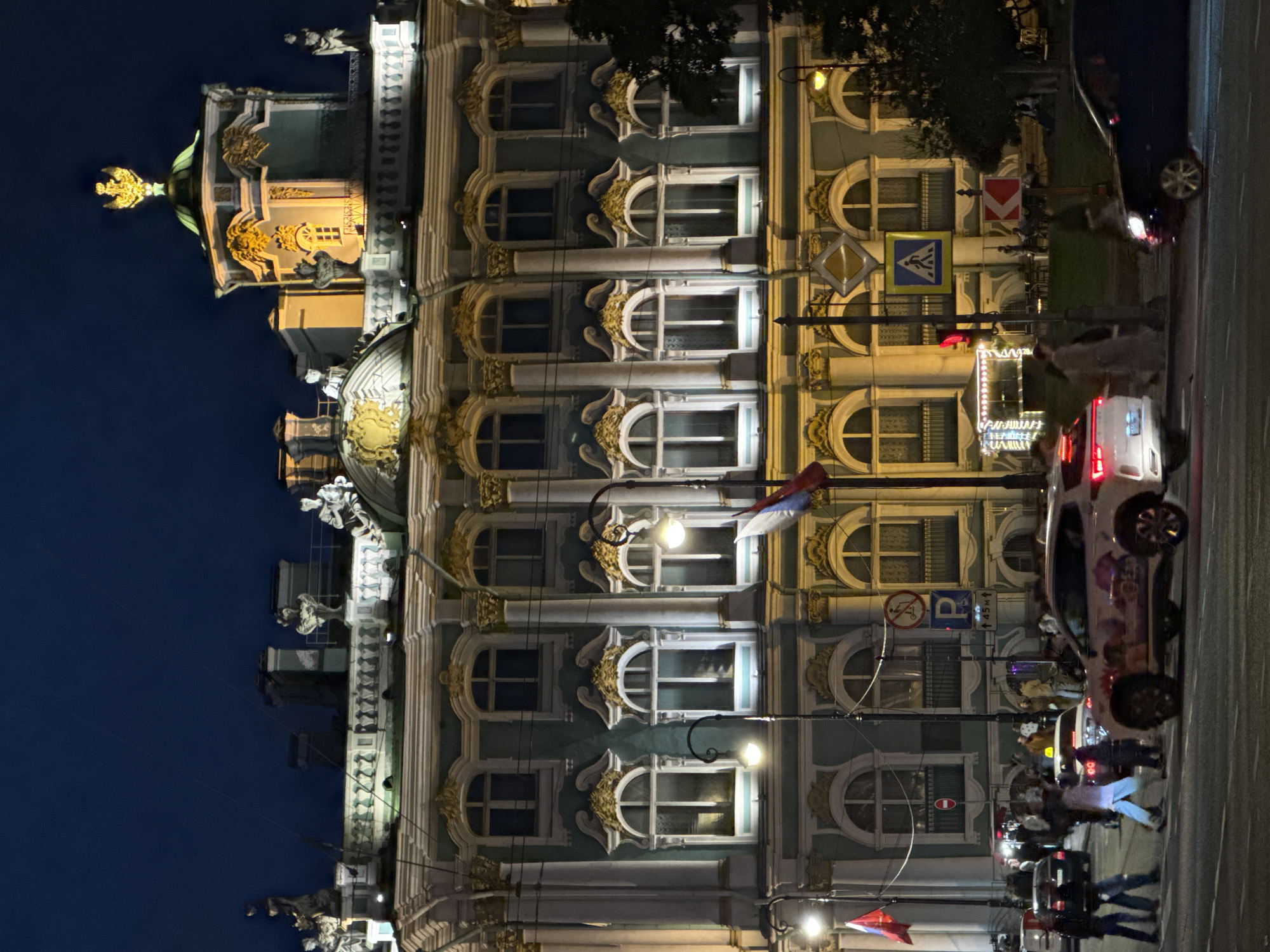 An outdoor, nighttime, eye-level, medium shot captures the grandeur of a large, ornate building in the city of St. Petersburg, Russia. The building, illuminated by artificial lights, showcases intricate architectural details, including statues adorning its roofline, arched windows with decorative gold trim, and classical columns. A prominent golden eagle emblem crowns a domed structure on the building's roof.
In the foreground, a street scene unfolds. Several cars, including a white SUV with colorful graphics and a dark sedan, are visible on a road, some in motion, suggesting traffic. Pedestrians are crossing the street in various directions, some illuminated by the streetlights and car headlights. Traffic signals and street signs, including a pedestrian crossing sign and a parking sign, are present. Russian flags are flown from poles. The overall atmosphere is one of a bustling urban environment at night, with the historic building serving as a striking backdrop.