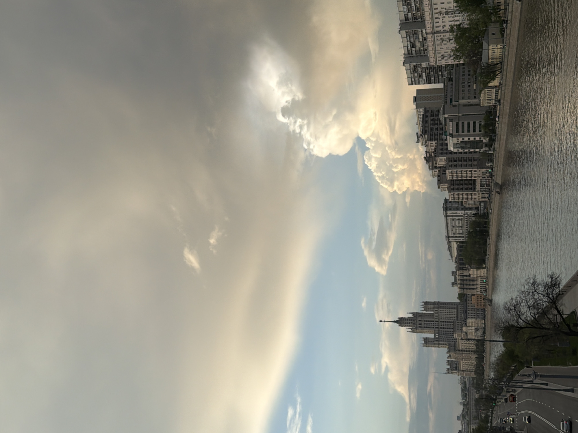 The image shows a cityscape with a river and cloudy sky. The buildings are tall and closely packed together, suggesting a dense urban environment. One prominent building has a spire-like structure on top. The river is on the right, and a road with cars is visible on the bottom right. The sky is filled with various cloud formations, some dark and ominous, and others bright and illuminated.