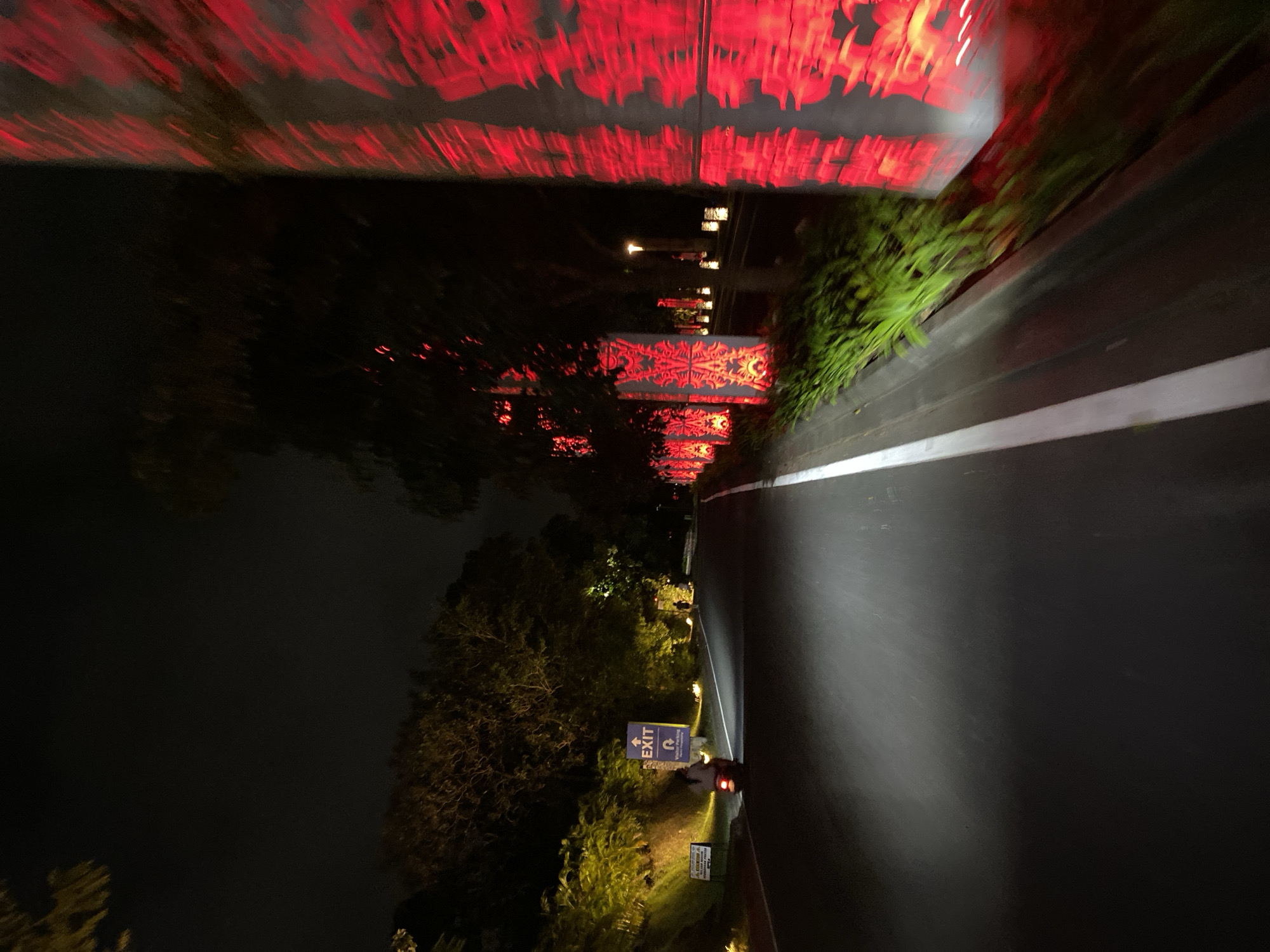 This image captures a nighttime view of a paved road in Kuta Selatan, Indonesia, possibly a resort or entertainment district, based on the decorative lighting. A single motorcycle with a rider is moving away from the camera on the left side of the road. To the left of the road, there's a slight incline with trees and bushes illuminated by warm yellow lights, and a blue "EXIT" sign with a logo is visible. On the right side of the road, there's a row of tall, slender structures adorned with vibrant red, patterned lighting, creating a dramatic visual effect against the dark backdrop. The motion blur indicates the photograph was likely taken from a moving vehicle or while the photographer was in motion. The overall atmosphere is illuminated by artificial lights, highlighting the road, surrounding vegetation, and the striking red decorations.