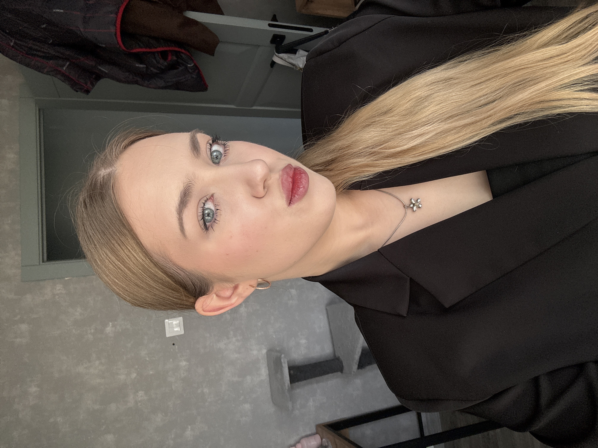 A young woman with blonde hair and blue eyes is seen in a close-up, portrait-style shot. She is wearing a black blazer and a delicate silver necklace with a flower-shaped pendant. She appears to be indoors, as indicated by the background featuring a textured grey wall, a door, and various furniture pieces. The image gives no indication that it was taken in the city of Chaikovsky, Russia.