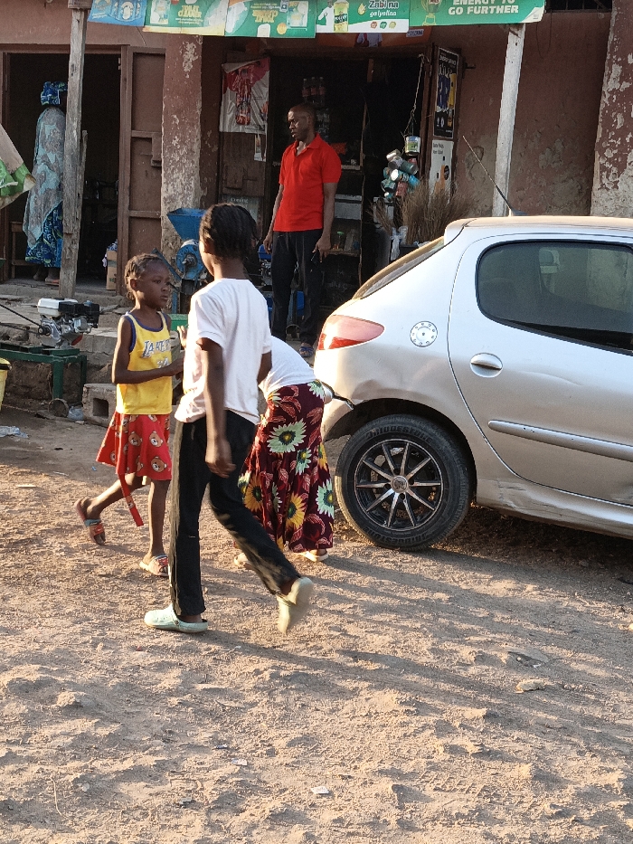 The scene depicts a street in Gwagwarwa, Nigeria, under natural daylight, likely late afternoon. The foreground features dusty, unpaved ground.
Five individuals are present. Centrally, two children are in motion or interaction. A child on the left, wearing a yellow sleeveless top with "LAKERS 32" and a red patterned skirt, faces right. Another child, with dark hair, wearing a white t-shirt and dark pants, walks right, passing the first child. An adult, partially obscured by a silver car, is seen bending at the waist near the car's rear, wearing a dark red skirt with prominent yellow and green floral patterns. In the background, an adult male wearing a red polo shirt and dark trousers stands in the entrance of a shop, holding an object in his right hand. Further inside the shop, an adult female in a light blue patterned dress is partially visible from behind.
A silver 3-door hatchback car, consistent with a Peugeot 206 model, is parked facing right, with its rear driver's side wheel featuring a black multi-spoke rim and "BRIDGESTONE" visible on the tire. The car shows a slight dent above the rear wheel arch.
The background comprises a single-story building, likely a commercial establishment, with reddish-brown walls and open doorways. A green banner with "ENERGY TO GO FURTHER" and other text is stretched across the top of the shop entrance. Various items, including brooms and containers, hang in the doorway. A small engine or piece of machinery on a green stand is visible on the far left.