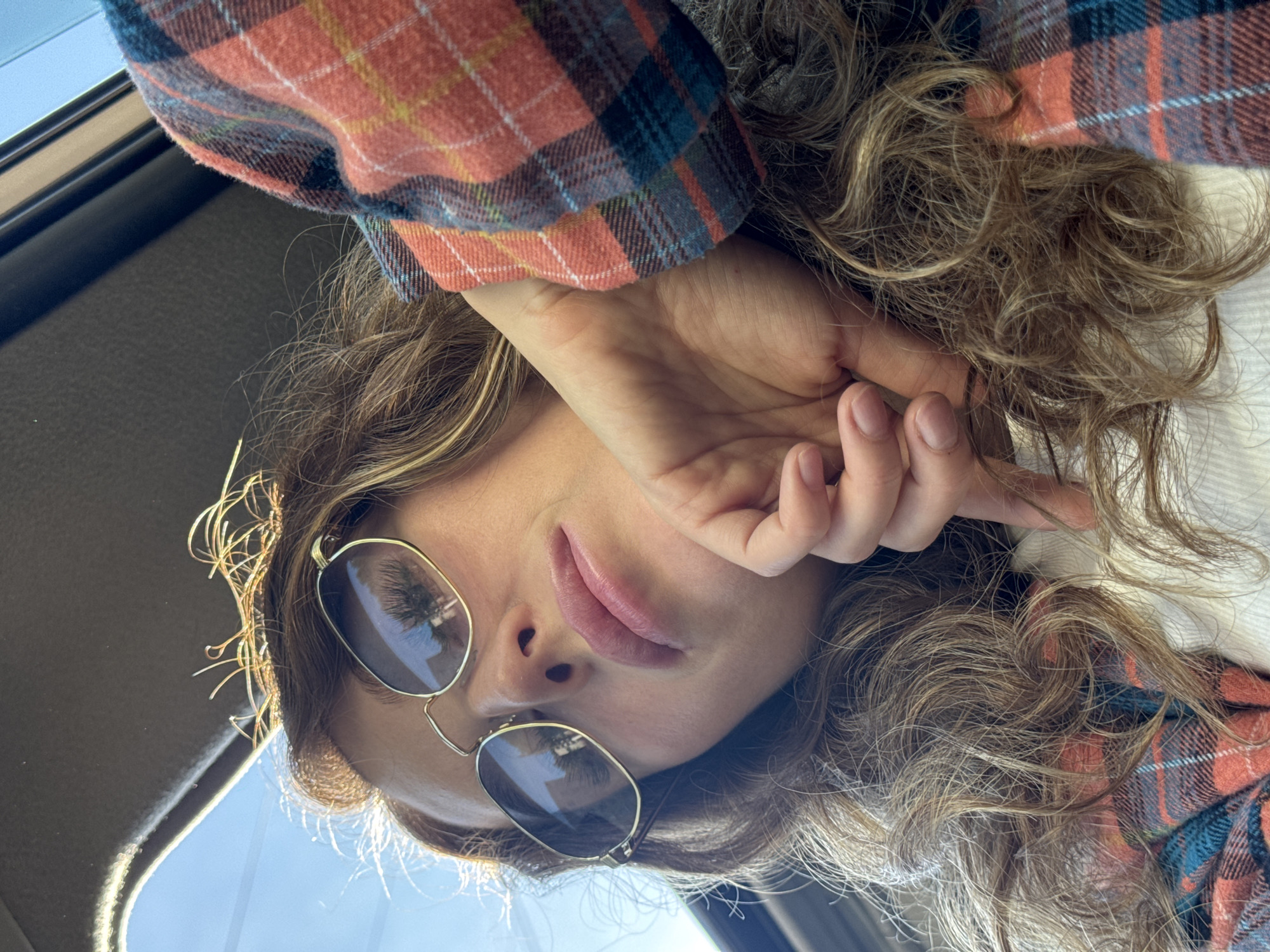 A woman with curly blonde hair and sunglasses is looking down at the camera. She is wearing a plaid shirt, and her hand is near her face. The light appears to be coming from outside. The location cannot be determined from this image.