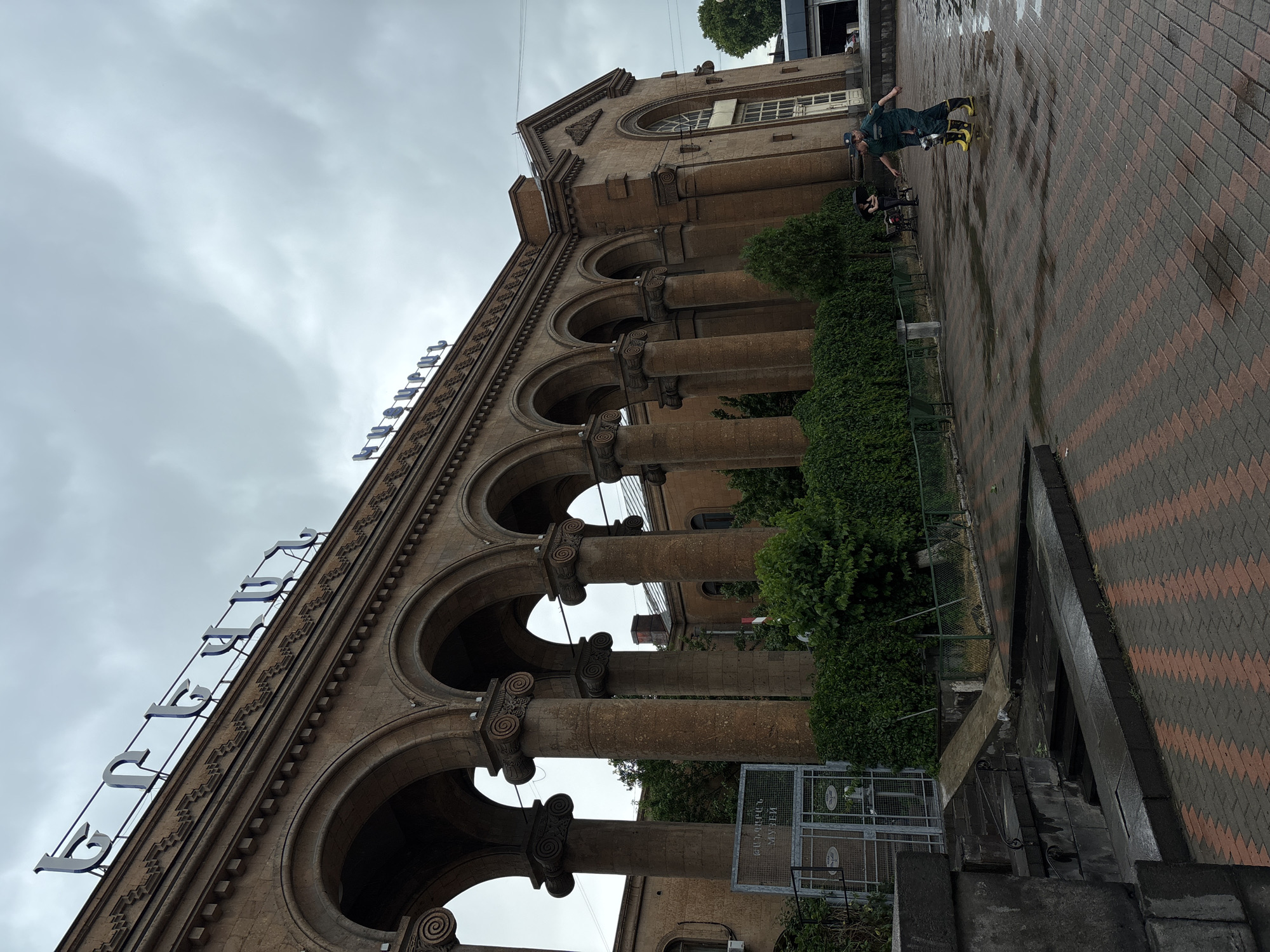 The image shows the exterior of the GUM (State Department Store) building in Yerevan, Armenia, a historic shopping arcade with a distinctive colonnade. The building's facade is made of reddish-brown stone, and it features a series of arched openings supported by columns. The top of the building displays the name "ԳՈՒՄ" in large, blue Armenian letters. The sky is overcast, suggesting a cloudy or rainy day. In the foreground, a person in a green uniform and yellow boots is holding a hose, possibly engaged in cleaning or maintenance. The ground is paved with a brick pattern, and there are wet patches, indicating recent rain. Lush green bushes and trees are visible behind the colonnade, contributing to the urban park-like setting. Several other individuals are present in the background, some standing under an umbrella, and others near a bench.