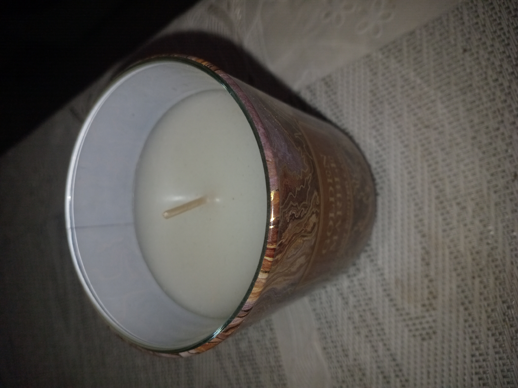 A lit candle in a decorative glass container sits on a textured surface. The candle itself is white and appears to be made of wax. The container has a patterned design in shades of gold and brown. The background is dark and out of focus, with some faint textures visible on the surface beneath the candle. The location in Perechyn, Ukraine is not discernible from the image.
